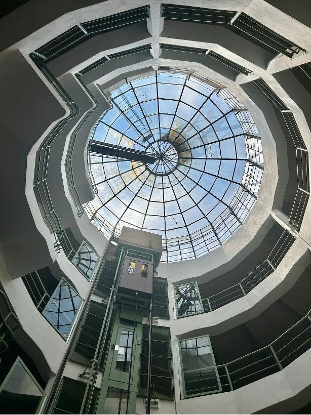 Blick nach oben in ein rundes Atrium mit gläserner Kuppel und Aufzug in der Mitte.