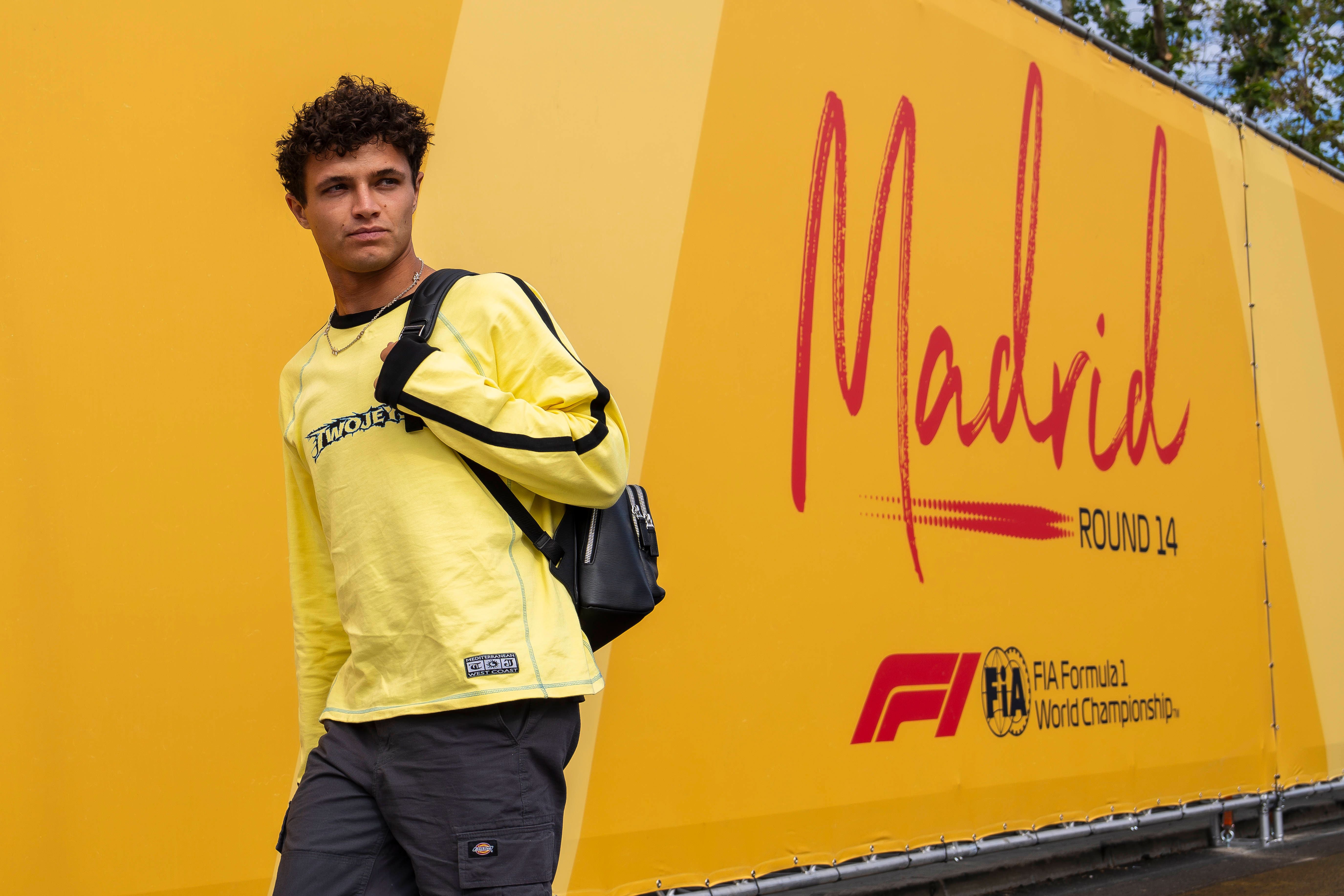 Lando Norris geht im gelben Langarmshirt mit Rucksack an einer gelben Wand mit dem Schriftzug Madrid vorbei.