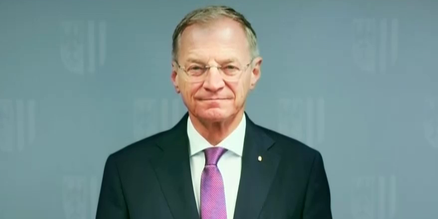 Ein Mann im Anzug mit lila Krawatte steht vor einem grauen Hintergrund.
