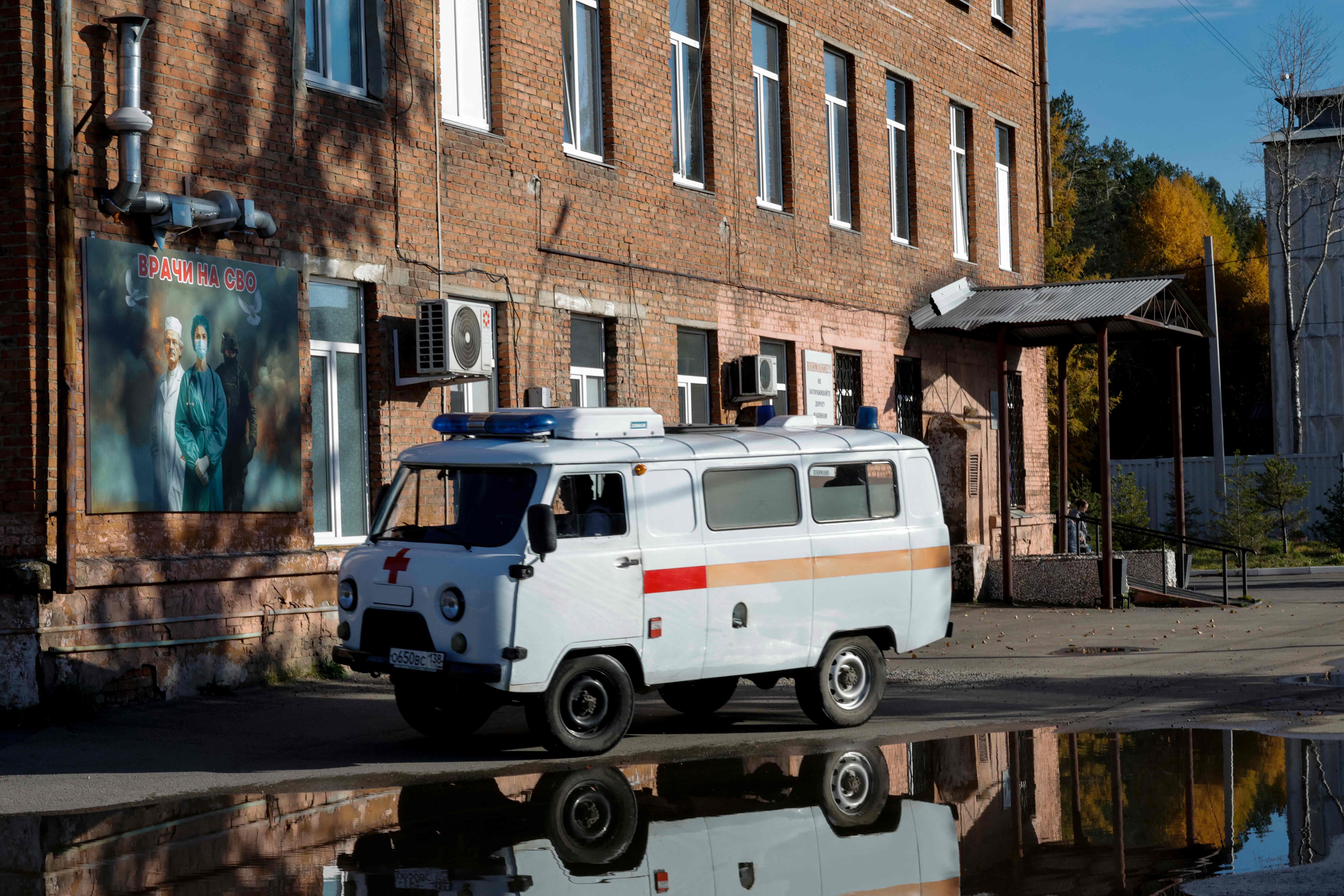 Ein Krankenwagen fährt an einem Krankenhausgebäude in Shelekhov vorbei, Spiegelung im Wasser sichtbar.