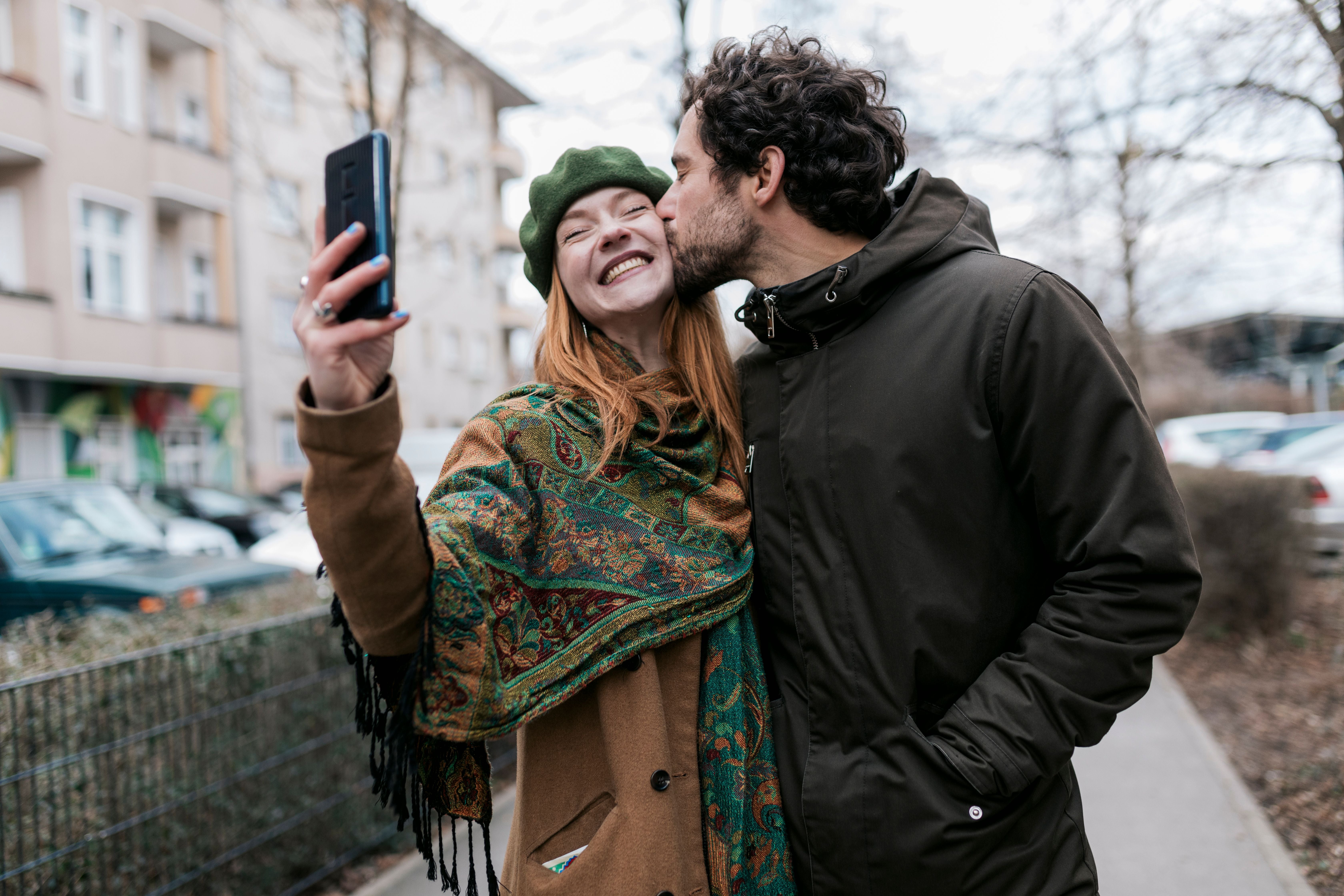 Ein Mann küsst seine Freundin auf die Wange, während sie ein Selfie in der Stadt macht.