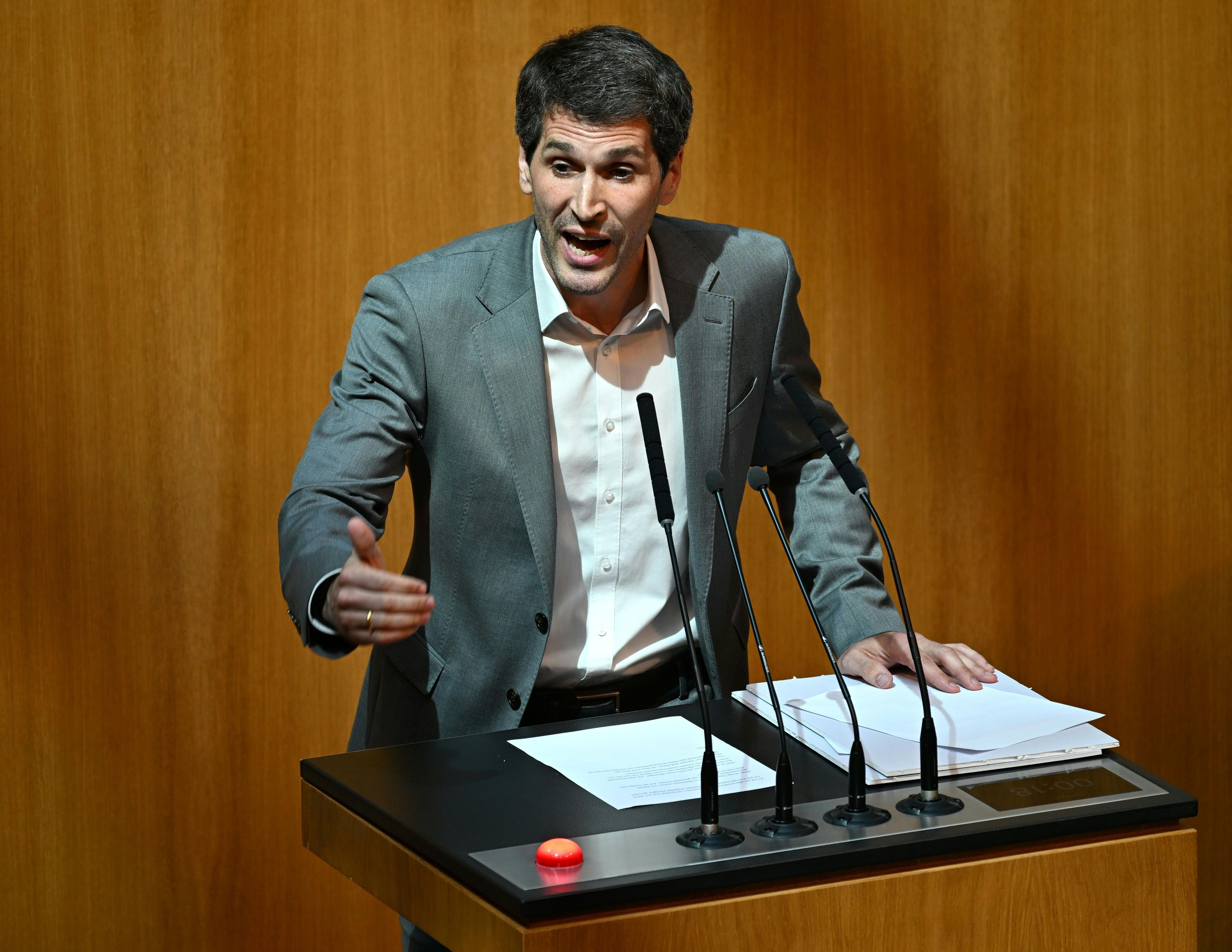 Ein Mann im grauen Anzug spricht gestikulierend an einem Rednerpult im Parlament.