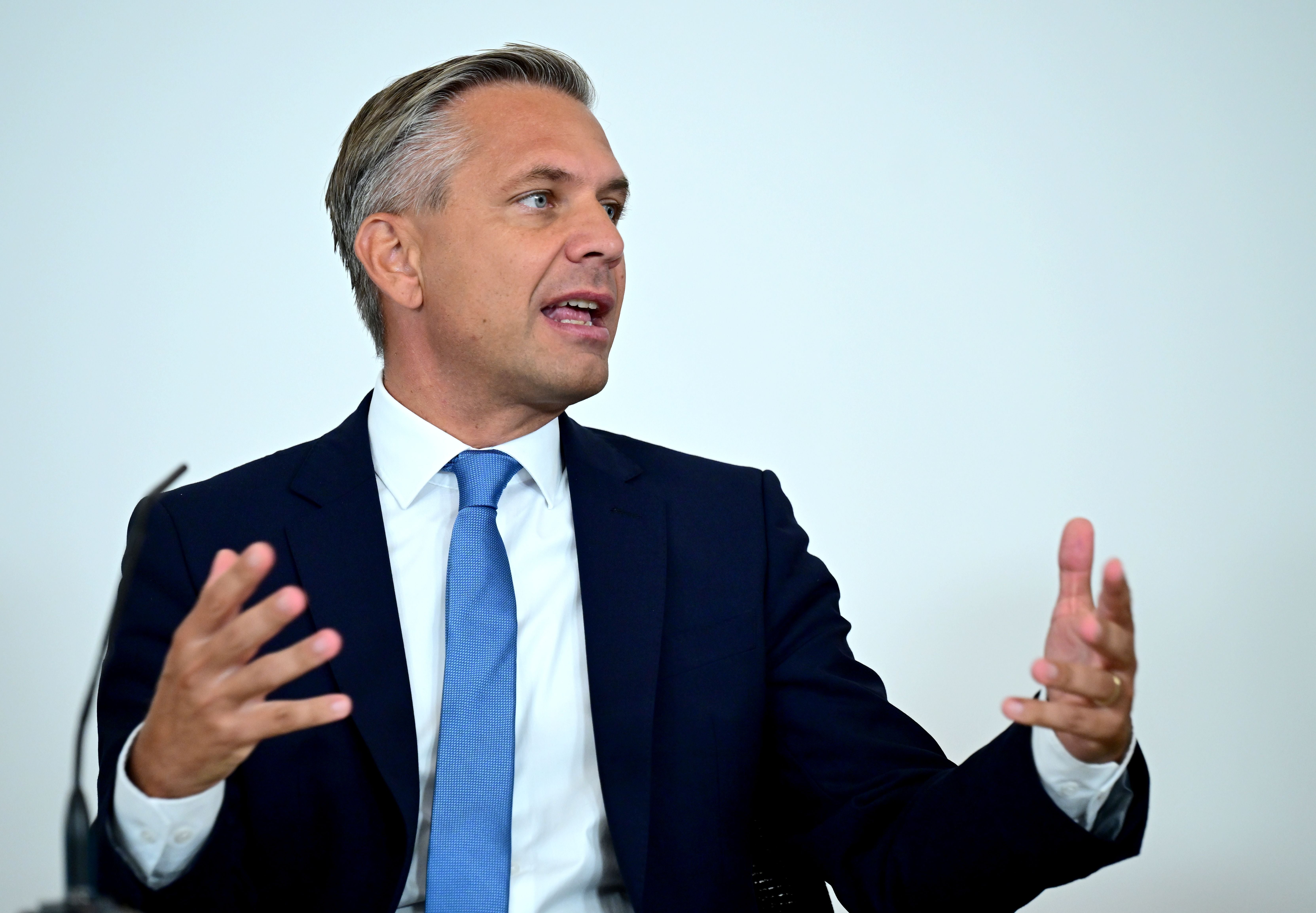 Ein Mann im Anzug spricht gestikulierend bei einer Pressekonferenz vor hellem Hintergrund.