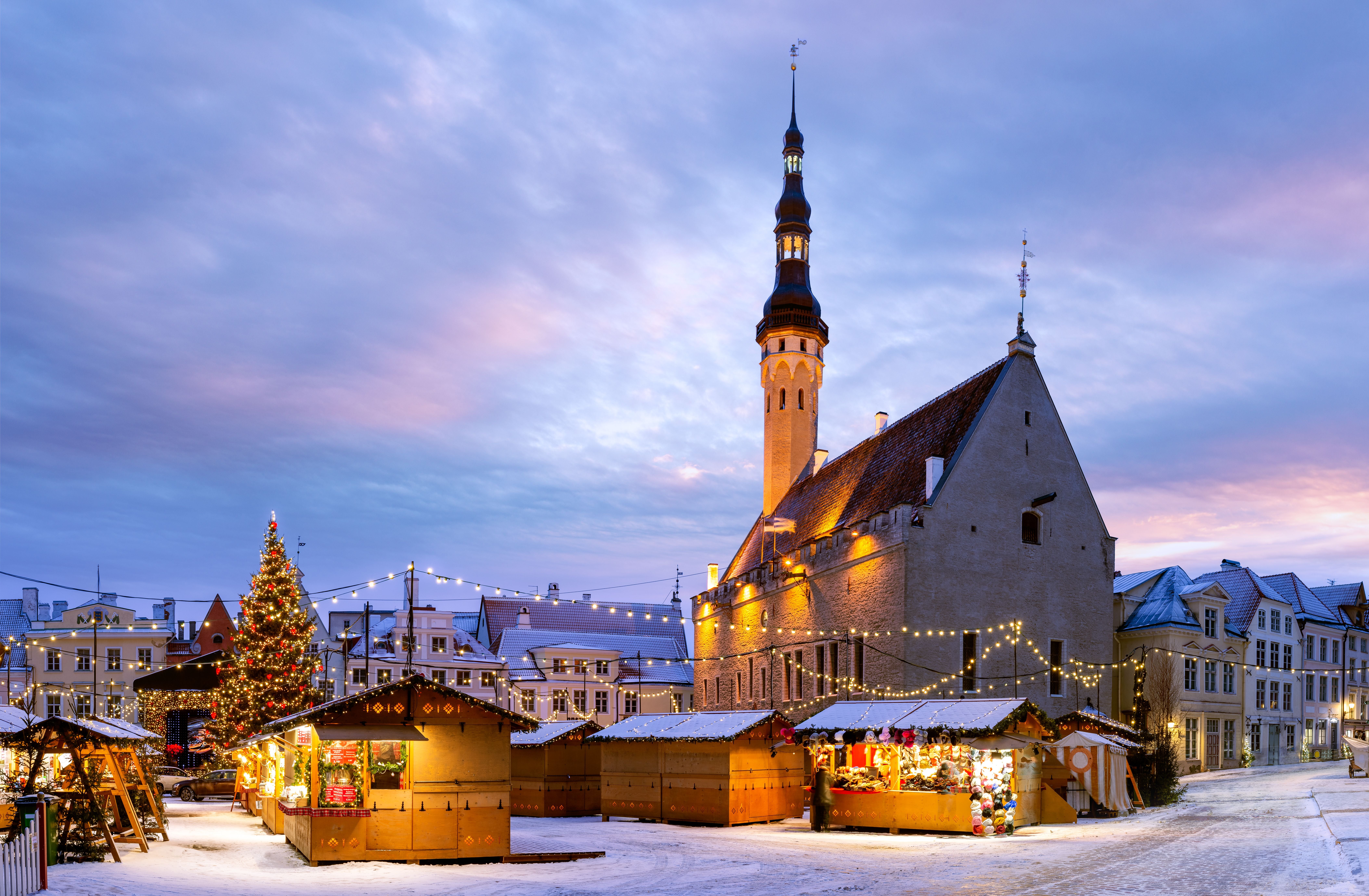 Weihnachtsmarkt mit beleuchtetem Rathaus und Weihnachtsbaum im verschneiten Tallinn bei Sonnenaufgang.