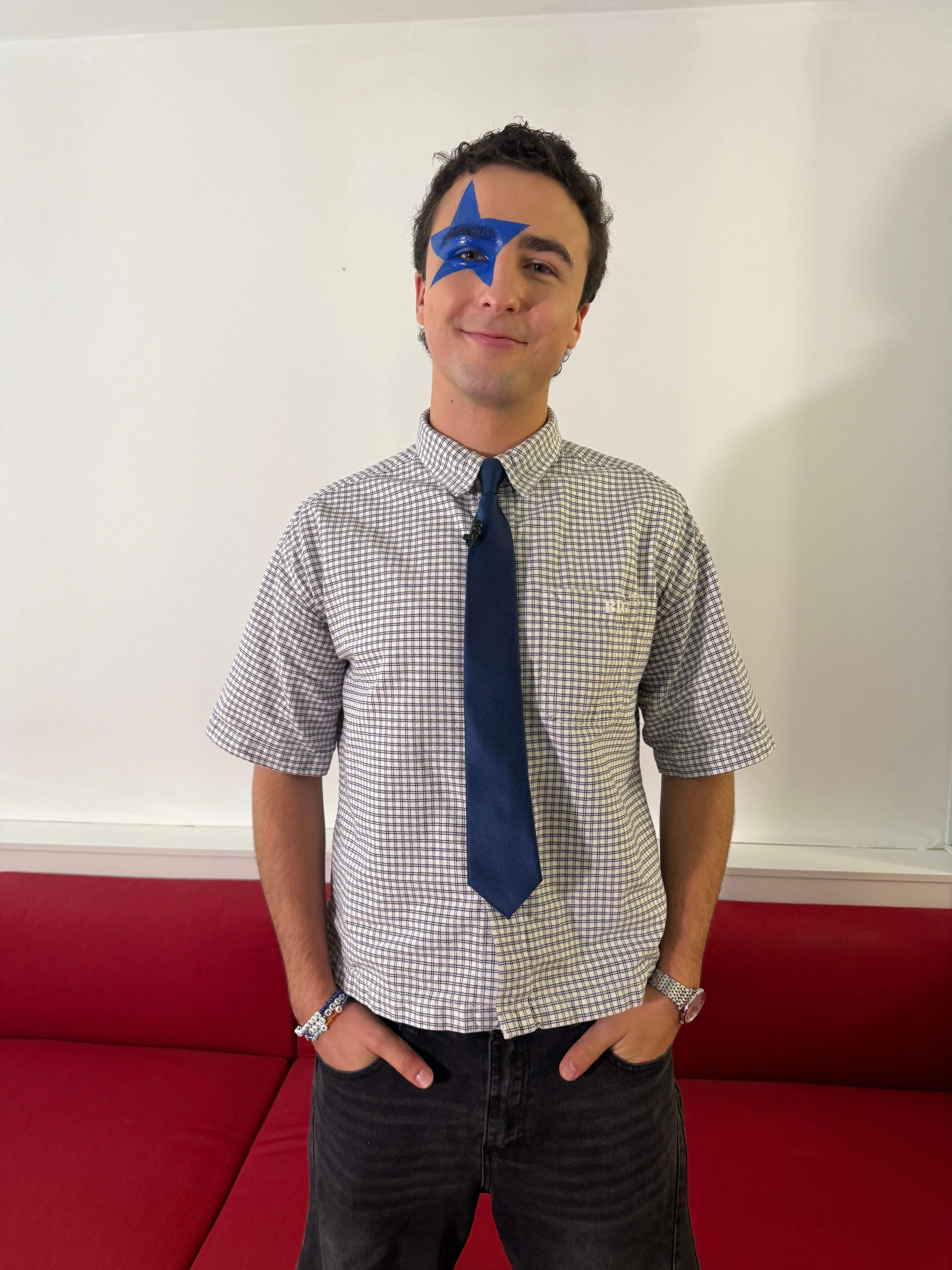 Junger Mann mit kariertem Hemd, blauer Krawatte und blauem Stern im Gesicht steht vor rotem Sofa.