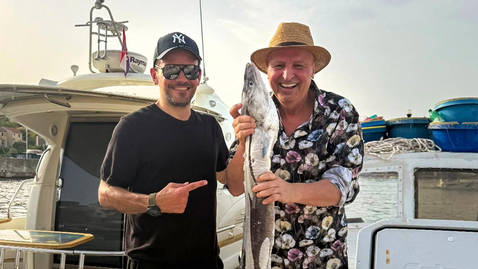 Zwei Männer stehen auf einem Boot, einer hält einen großen Fisch und beide lächeln.