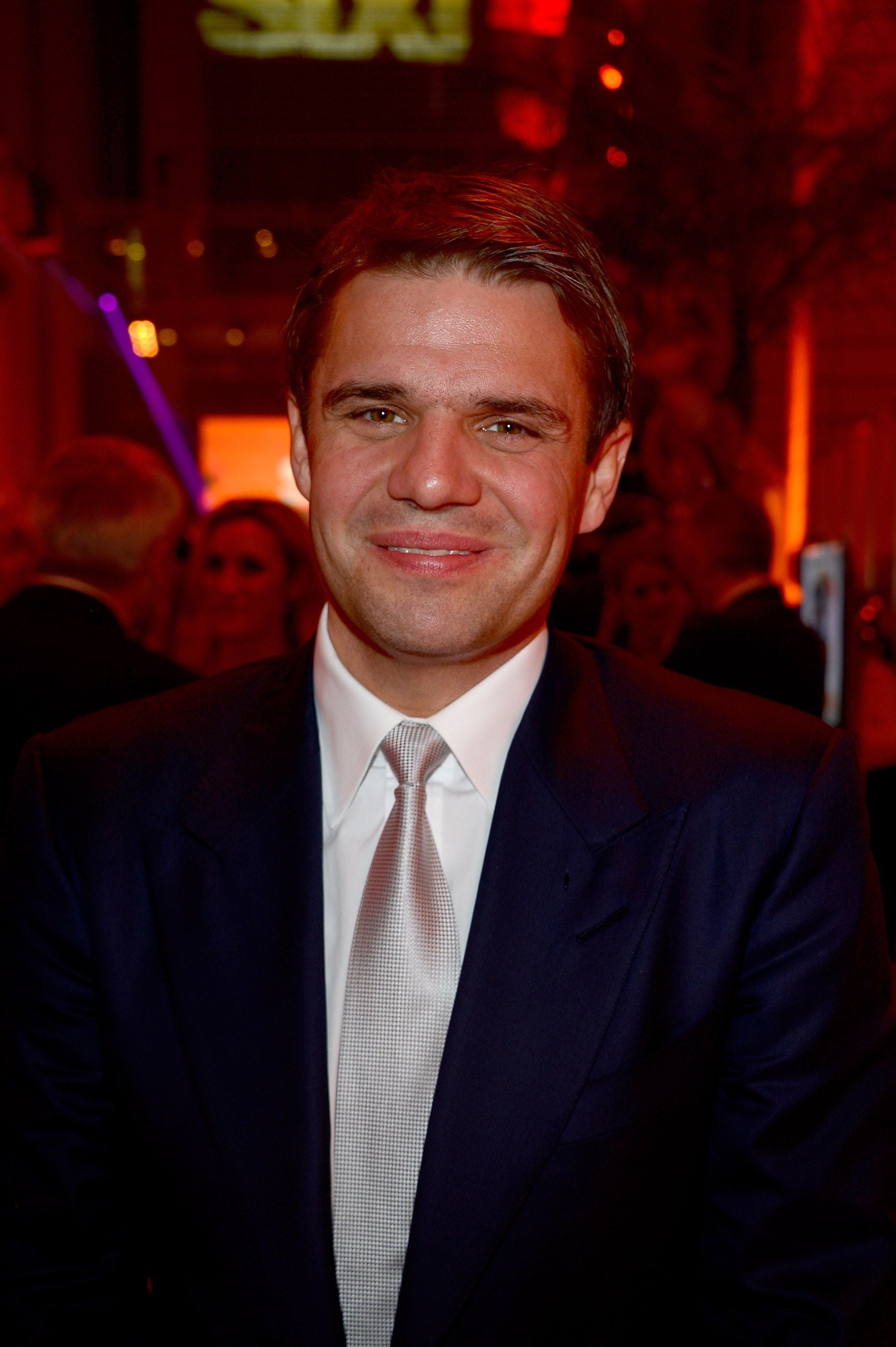 Ein Mann im Anzug lächelt auf einer Party mit rotem Licht im Hintergrund in die Kamera.