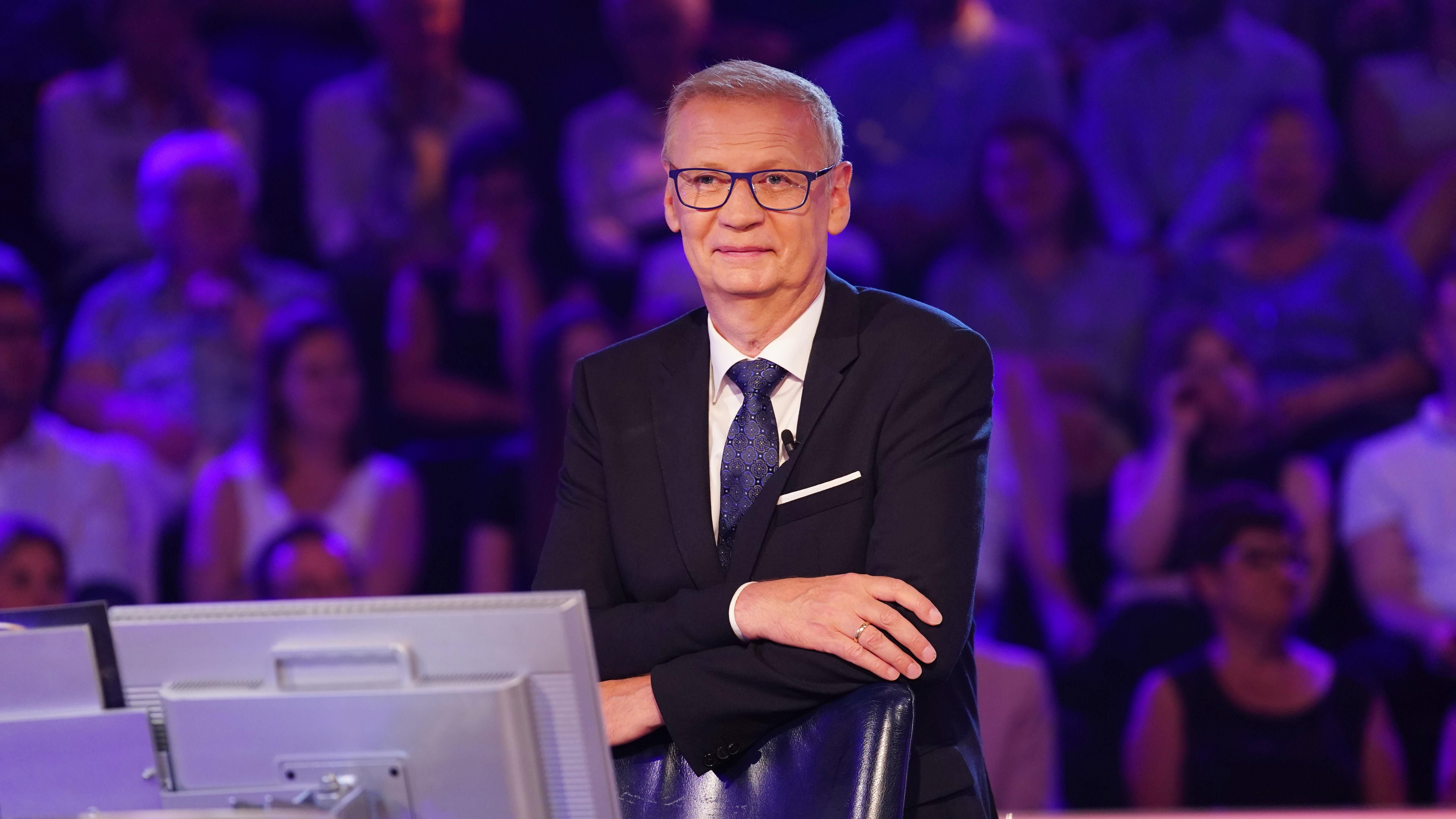 Ein Mann im Anzug steht im Fernsehstudio vor Monitoren, Publikum sitzt im Hintergrund.