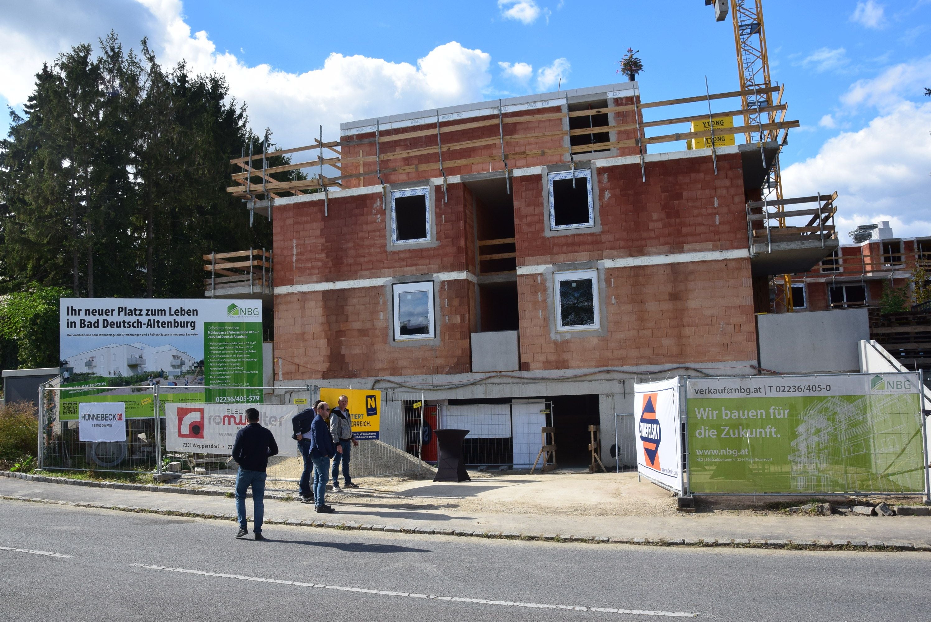 Mehrstöckiges Wohnhaus im Bau mit Bauarbeitern und Werbeschildern davor.