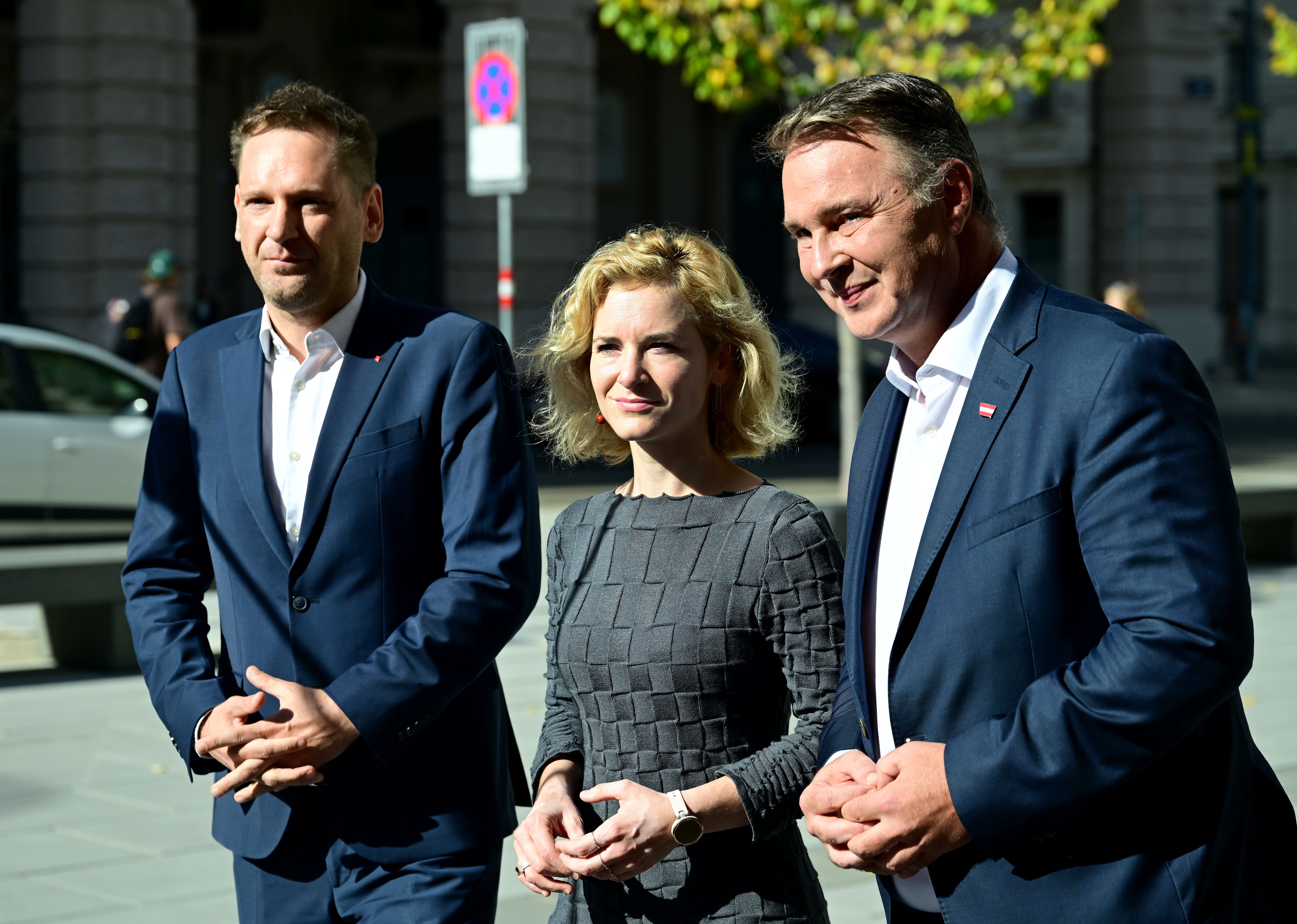 Drei Personen im Business-Outfit stehen draußen, lächeln und schauen in dieselbe Richtung.
