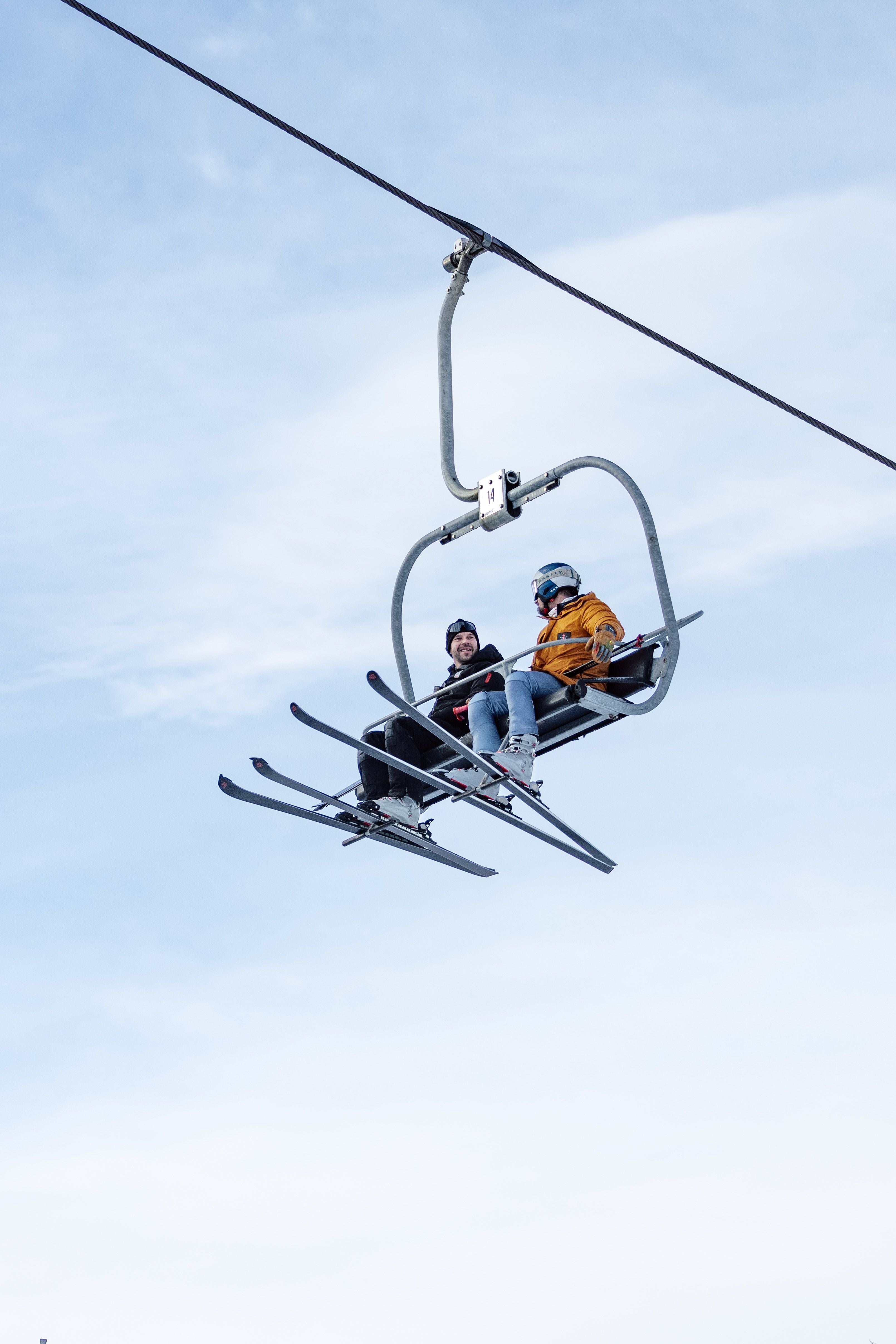 Zwei Skifahrer sitzen auf einem Sessellift vor blauem Himmel.