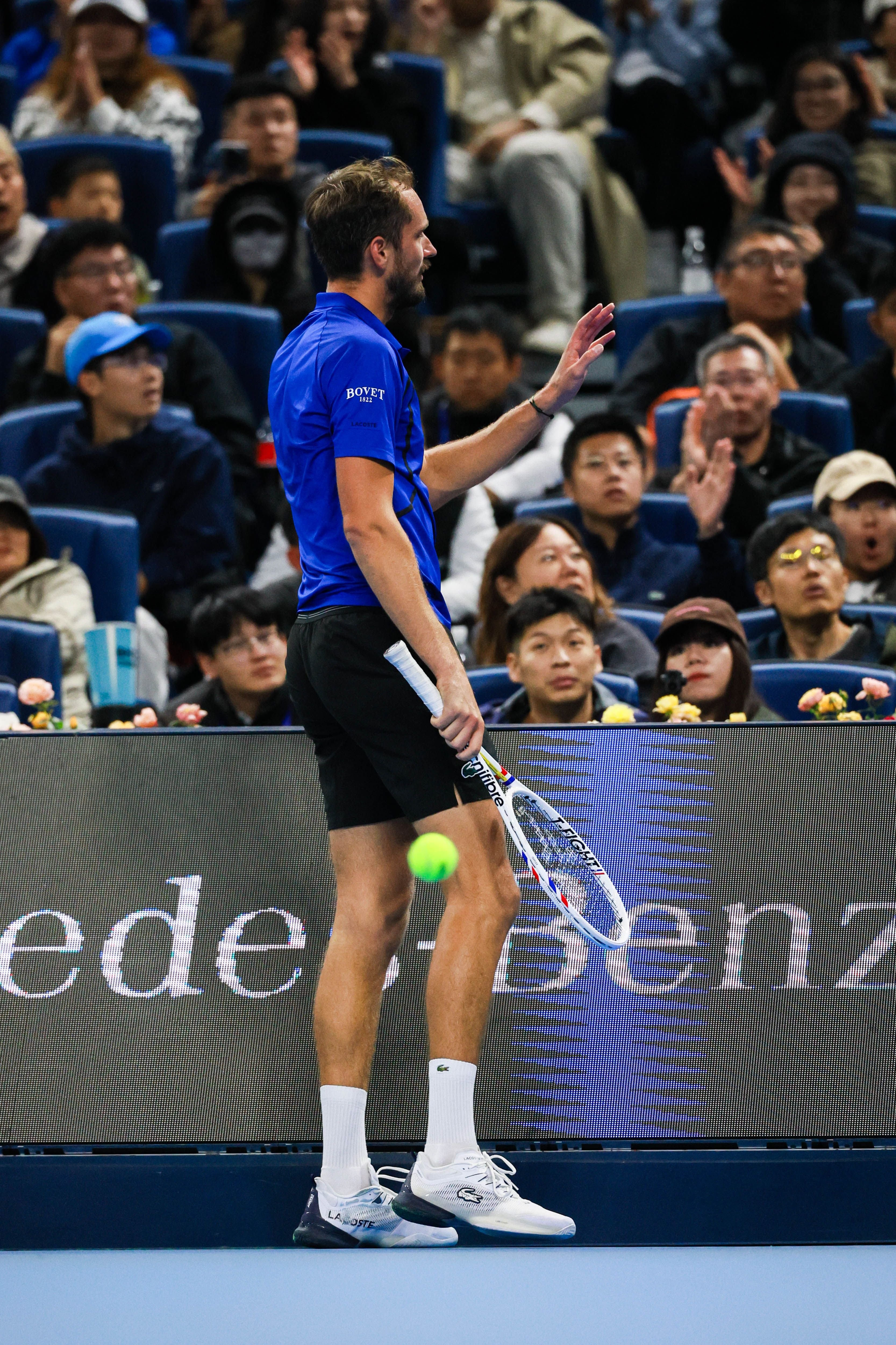 Ein Tennisspieler in blauer Kleidung steht auf dem Platz vor einem Publikum, Ball in der Luft.