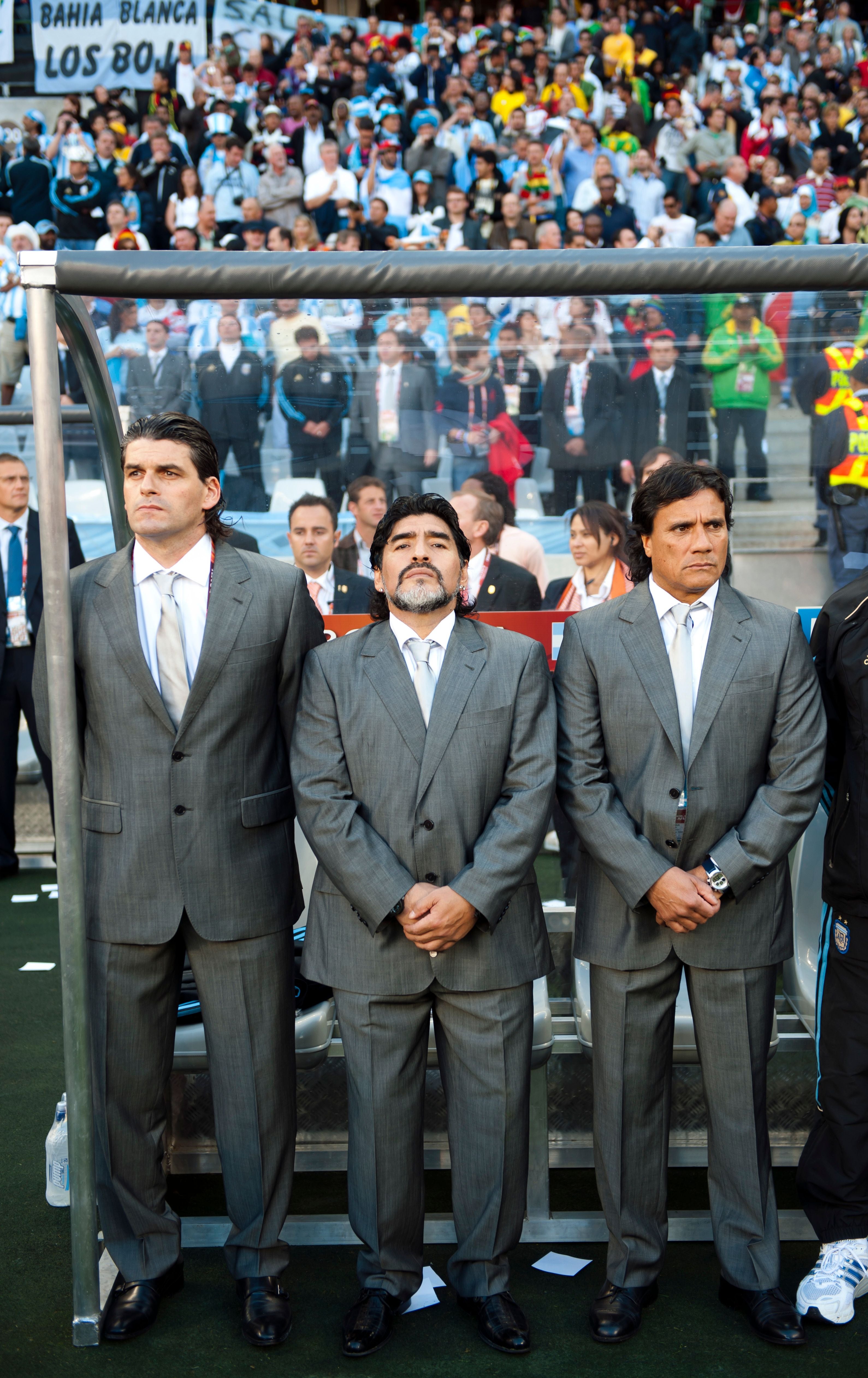 Diego Maradona und Alejandro Mancuso stehen nebeneinander in grauem Anzug und Krawatte am Spielfeldrand.