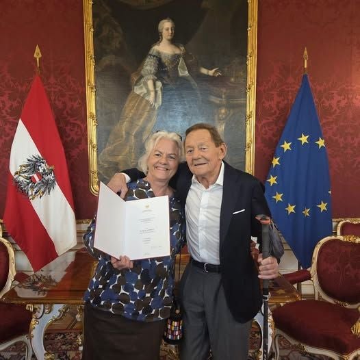Ein älteres Paar steht lächelnd vor österreichischer und EU-Flagge mit einer Urkunde in der Hand.