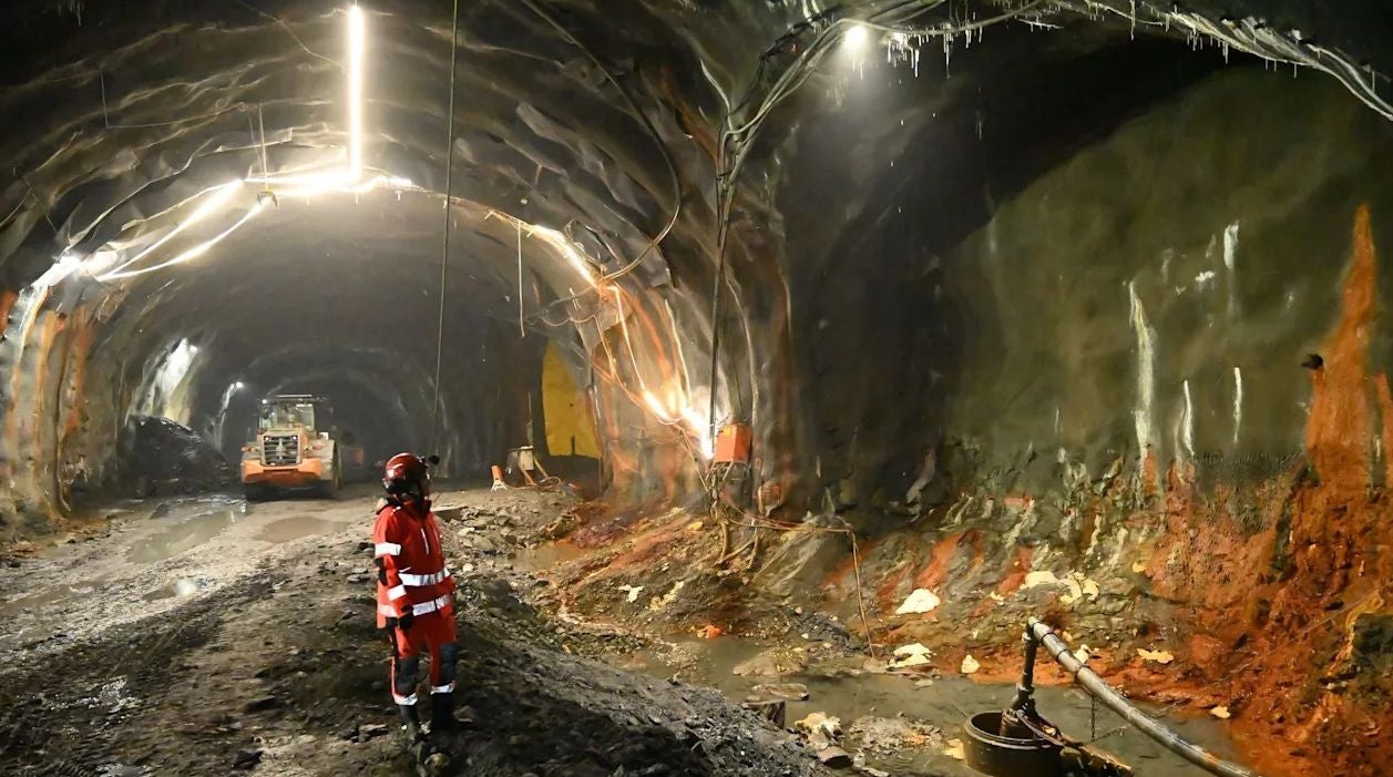 Arbeiter in Sicherheitskleidung steht in beleuchtetem Tunnel mit Bagger und feuchtem Boden.