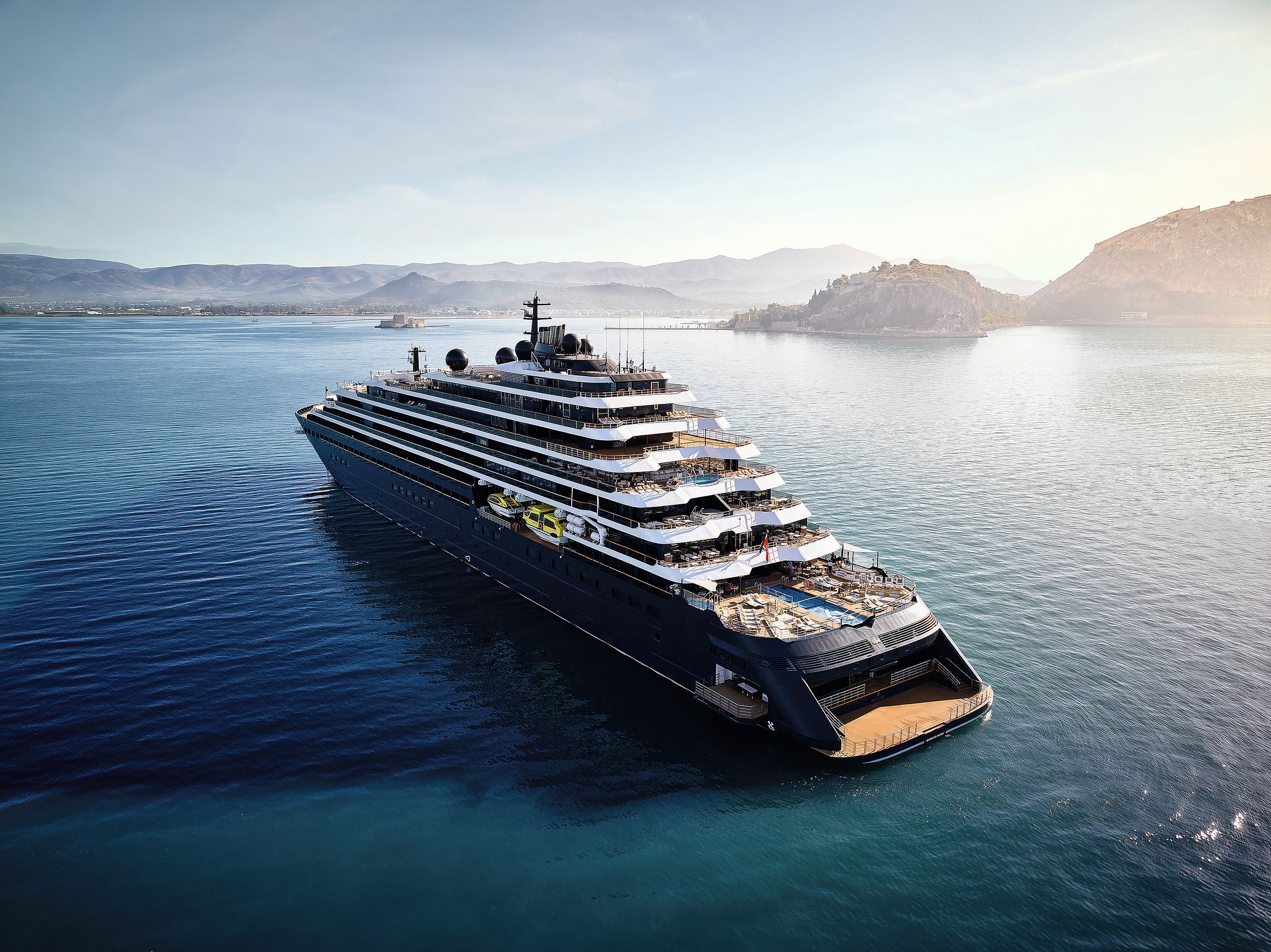Luxuriöses Kreuzfahrtschiff fährt auf ruhigem Meer, im Hintergrund Berge und Küste.