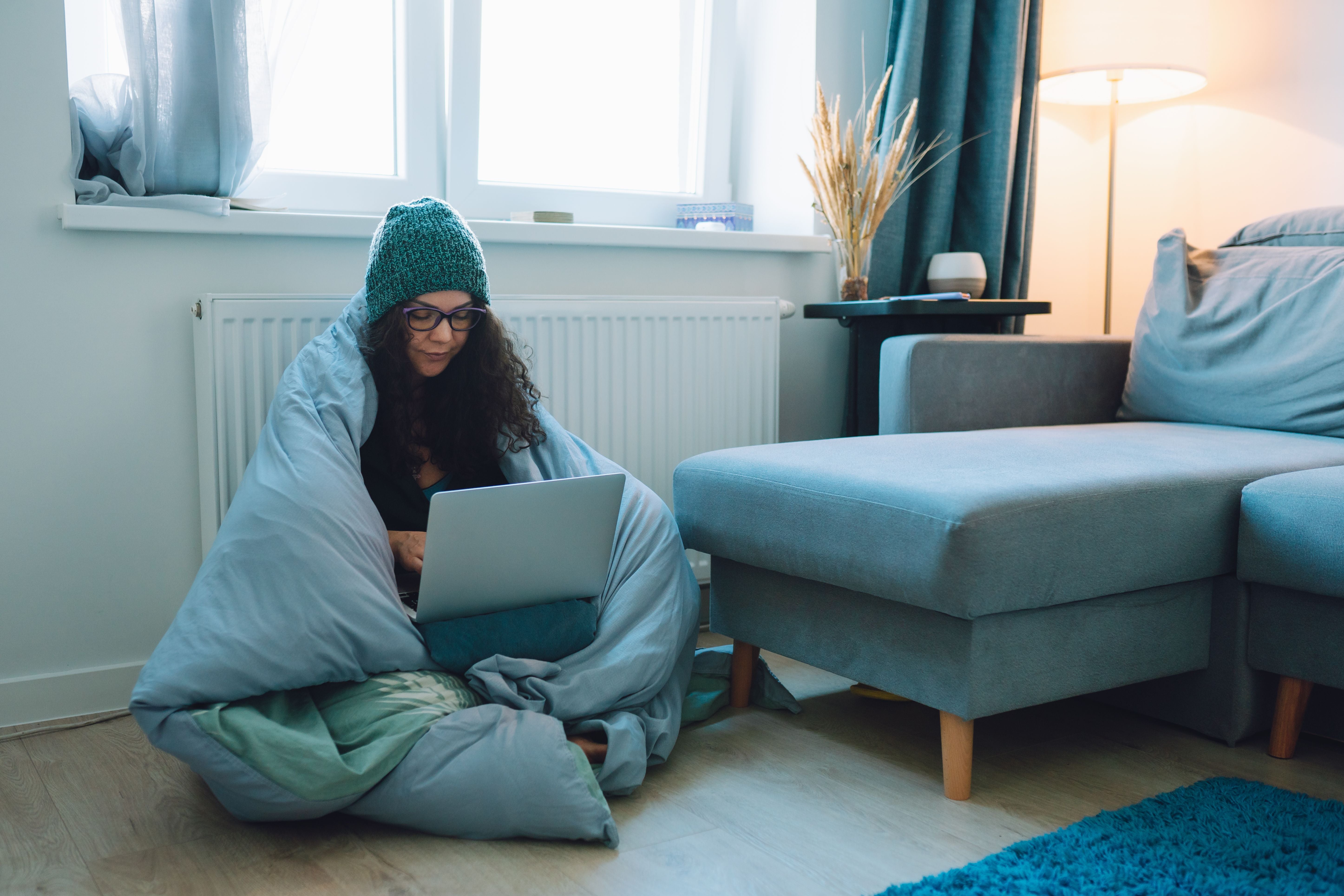 Frau sitzt eingewickelt in eine Decke mit Laptop neben der Heizung und trägt eine Wollmütze.