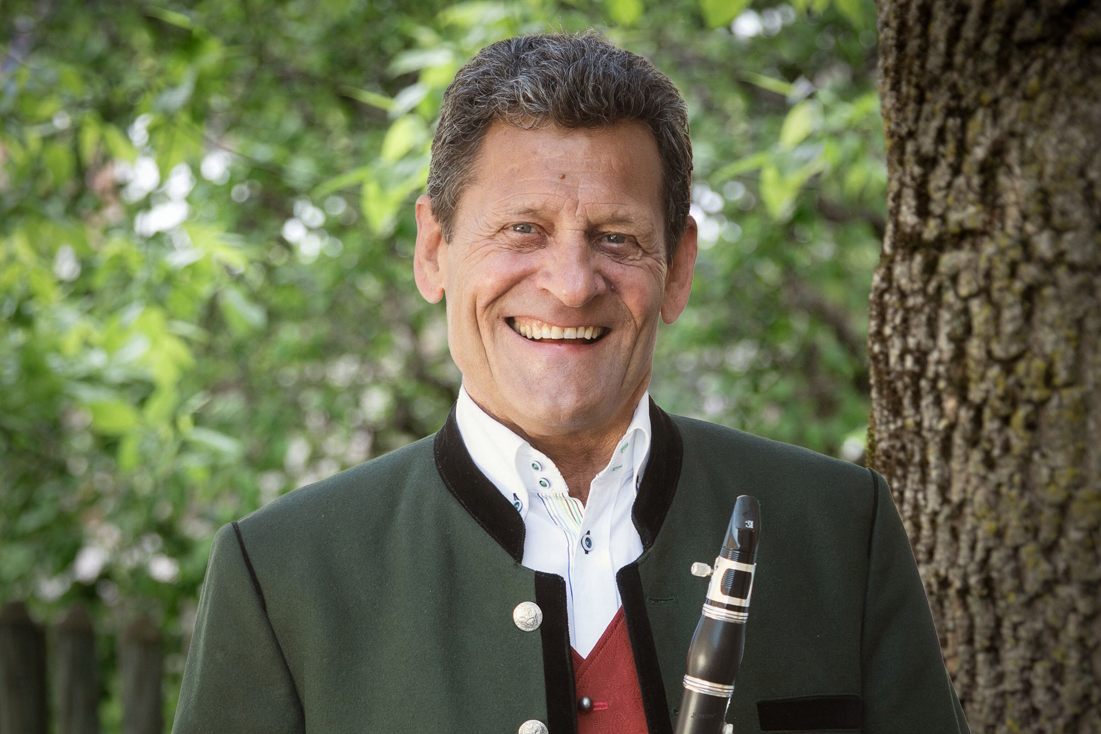 Ein lachender Mann in Tracht hält eine Klarinette, im Hintergrund sind grüne Blätter zu sehen.