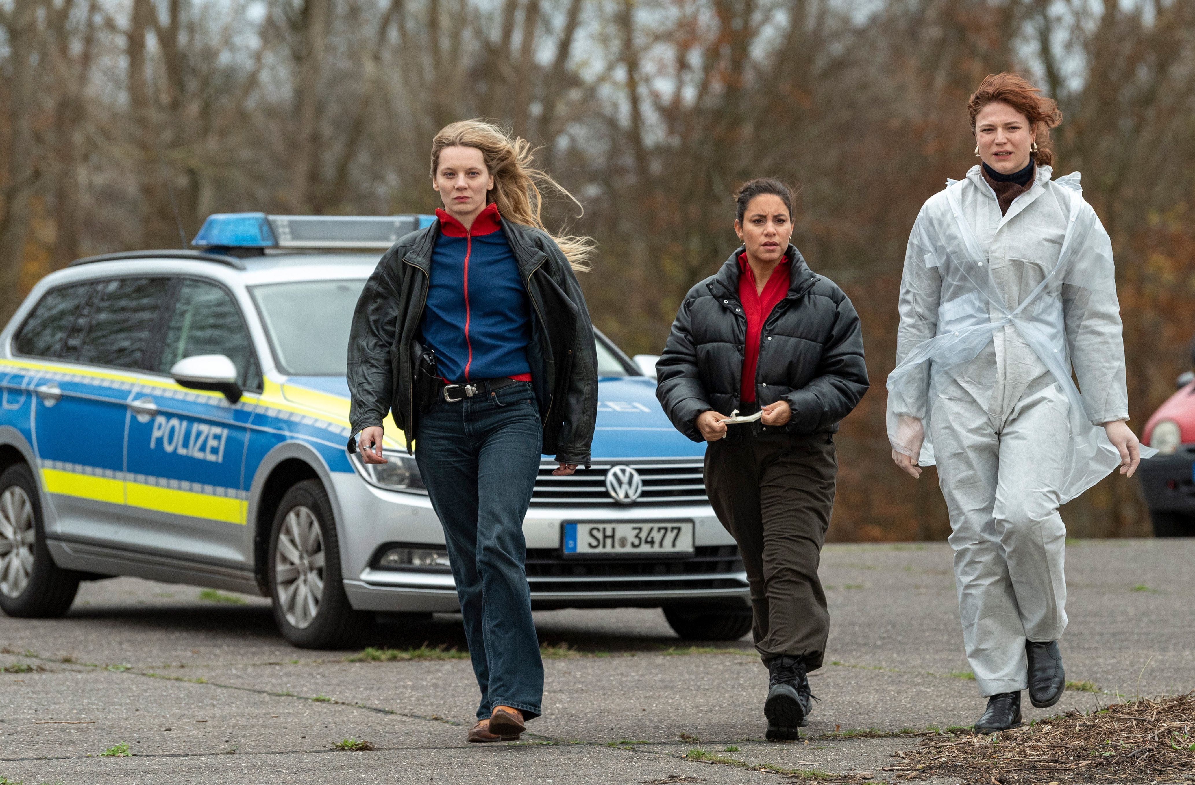 Drei Frauen gehen vor einem Polizeiauto, eine trägt einen weißen Forensik-Overall.