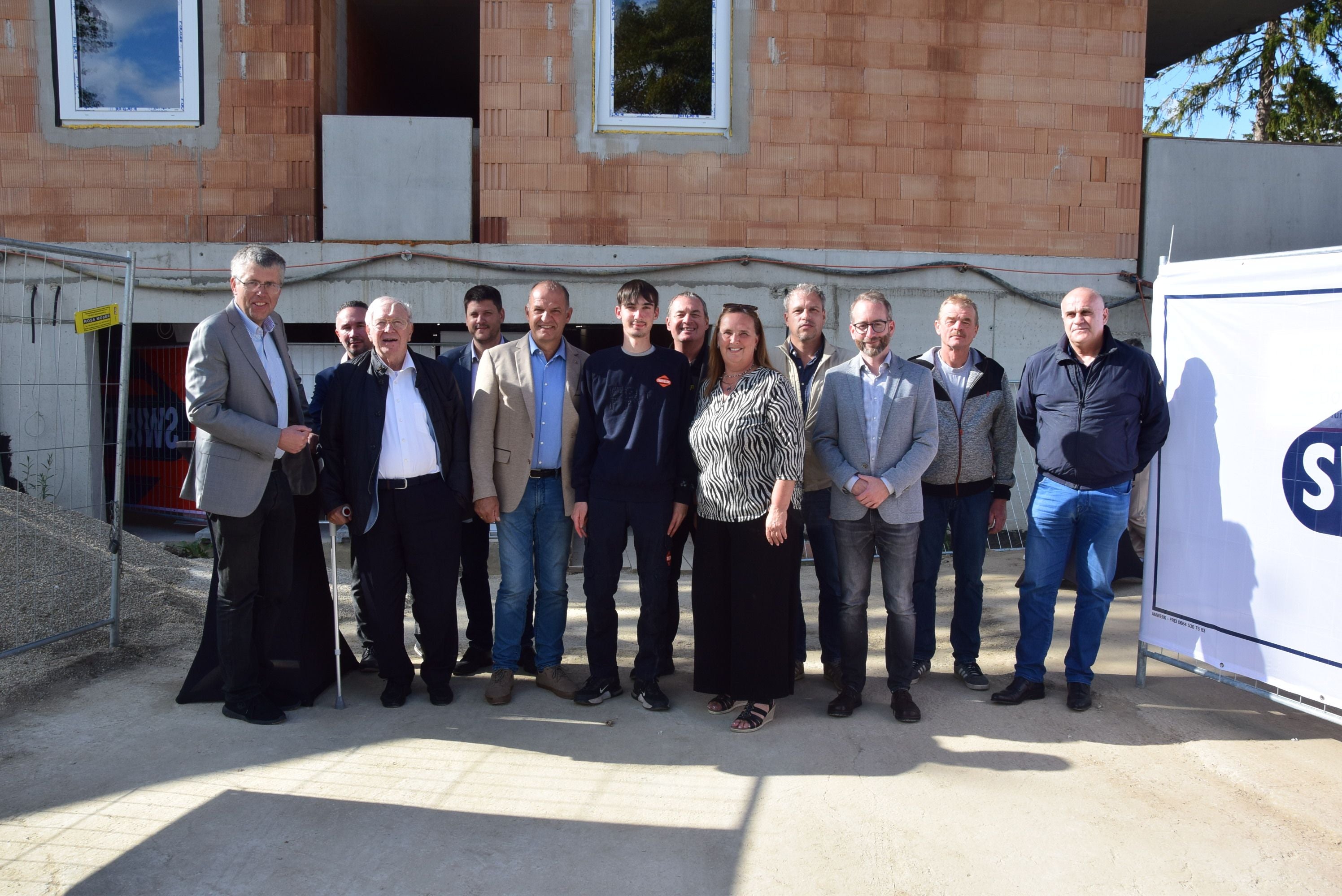 Eine Gruppe von Menschen posiert lächelnd vor einer Baustelle im Sonnenlicht.