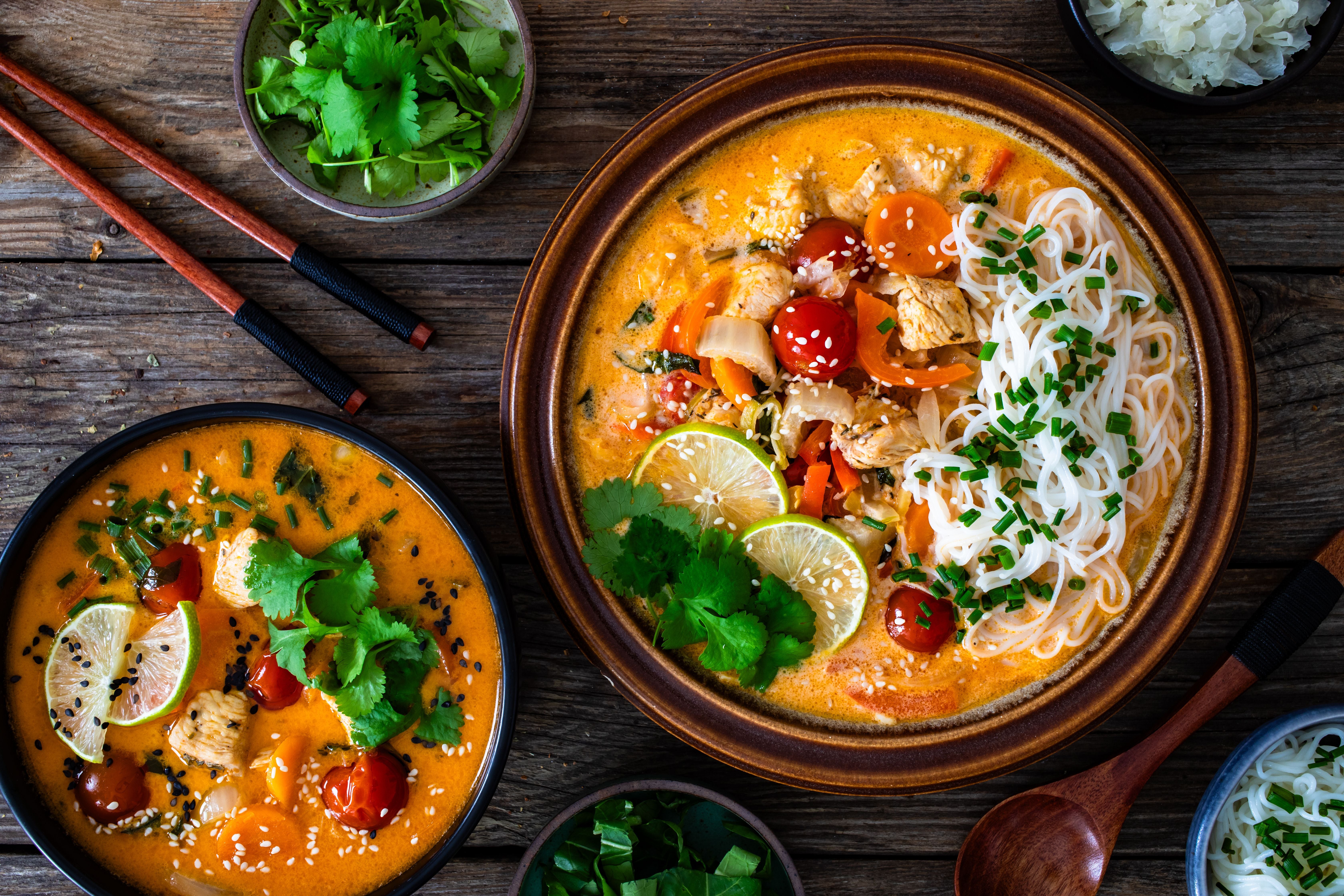 Tom Yum Suppe mit Hähnchen, Reisnudeln, Limetten und Koriander auf Holztisch angerichtet.