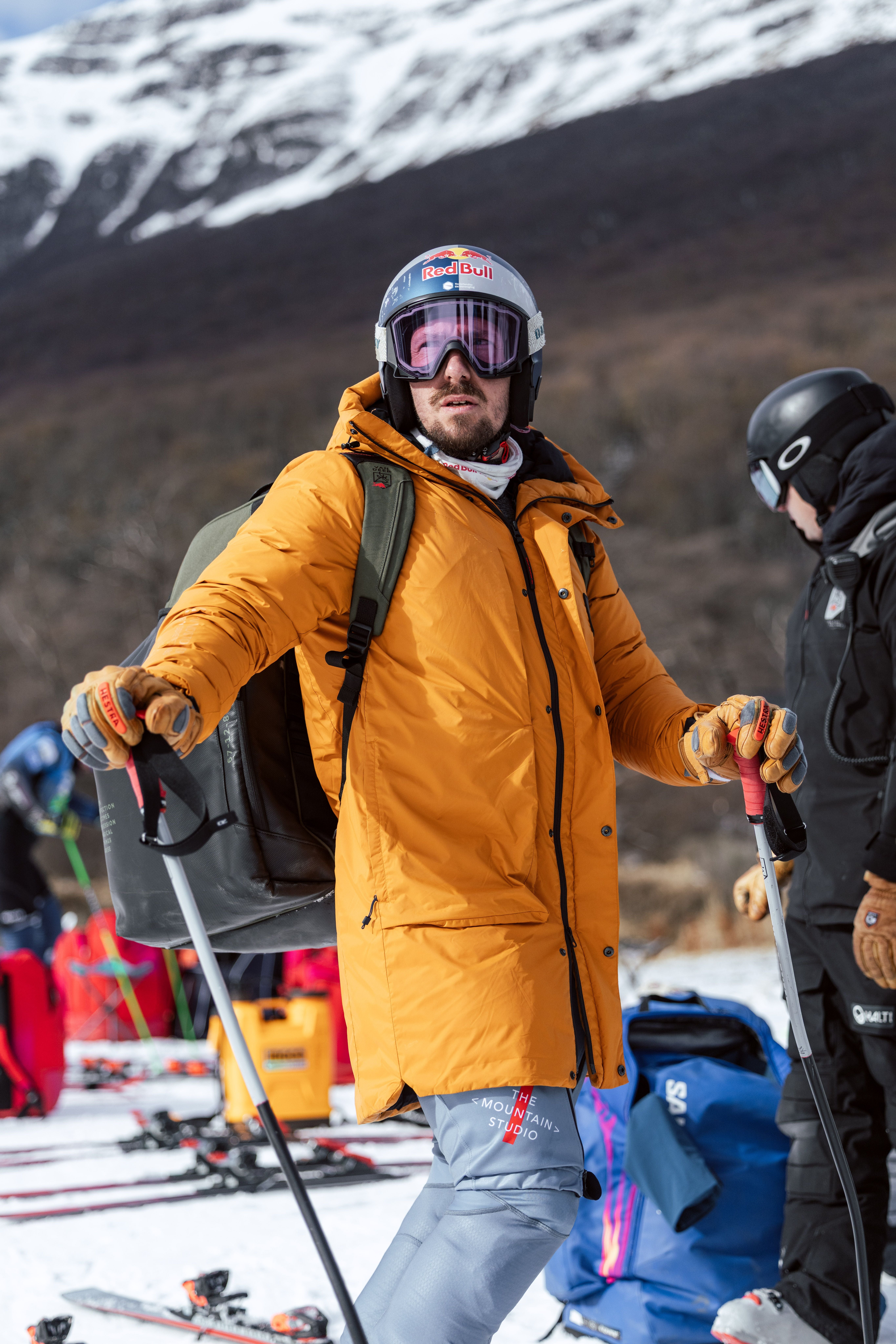 Ein Skifahrer in orangefarbener Jacke und Helm steht mit Skistöcken im Schnee vor Bergen.