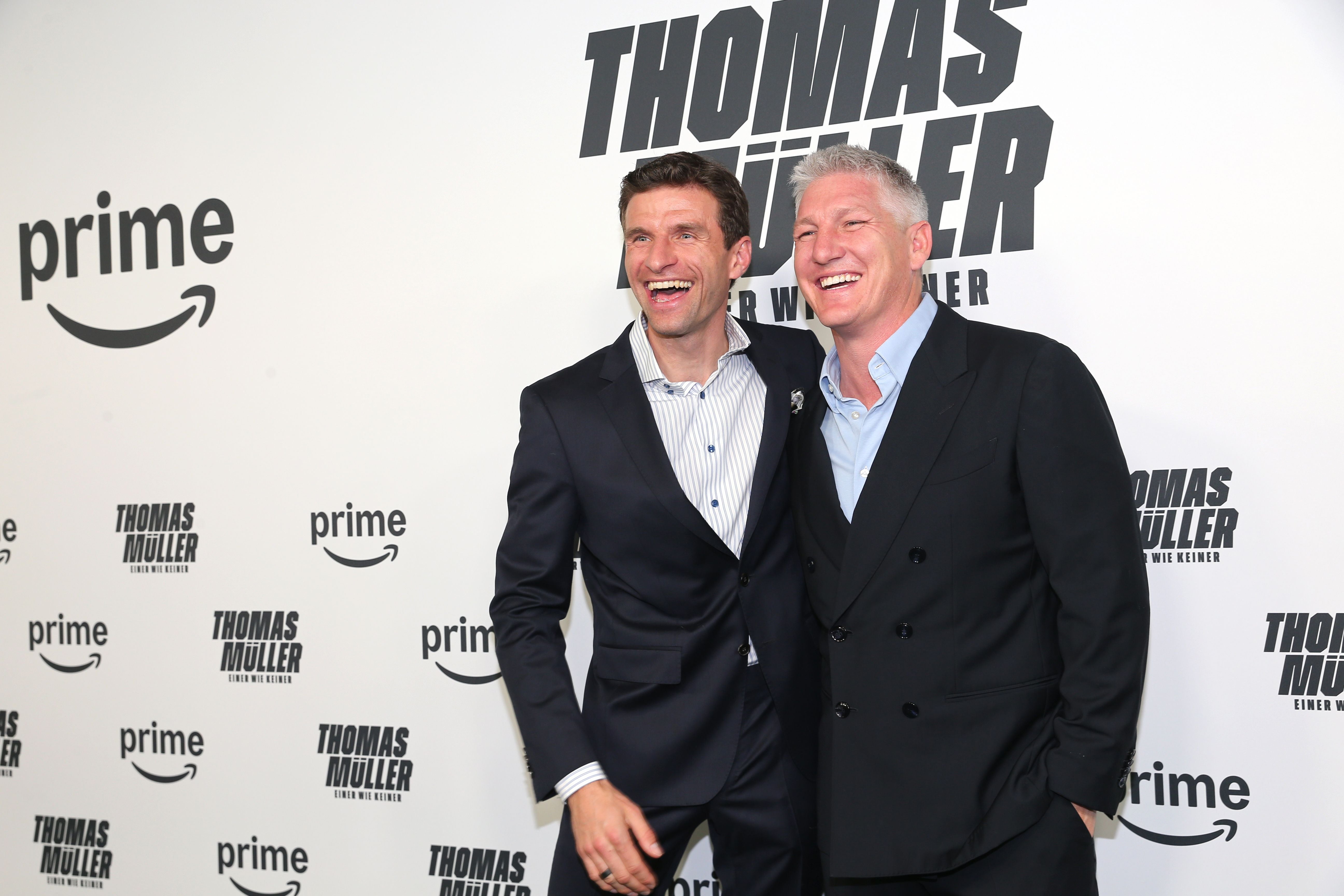 Zwei Männer im Anzug lachen auf einem roten Teppich vor einer Fotowand mit „Thomas Müller“-Schriftzug.