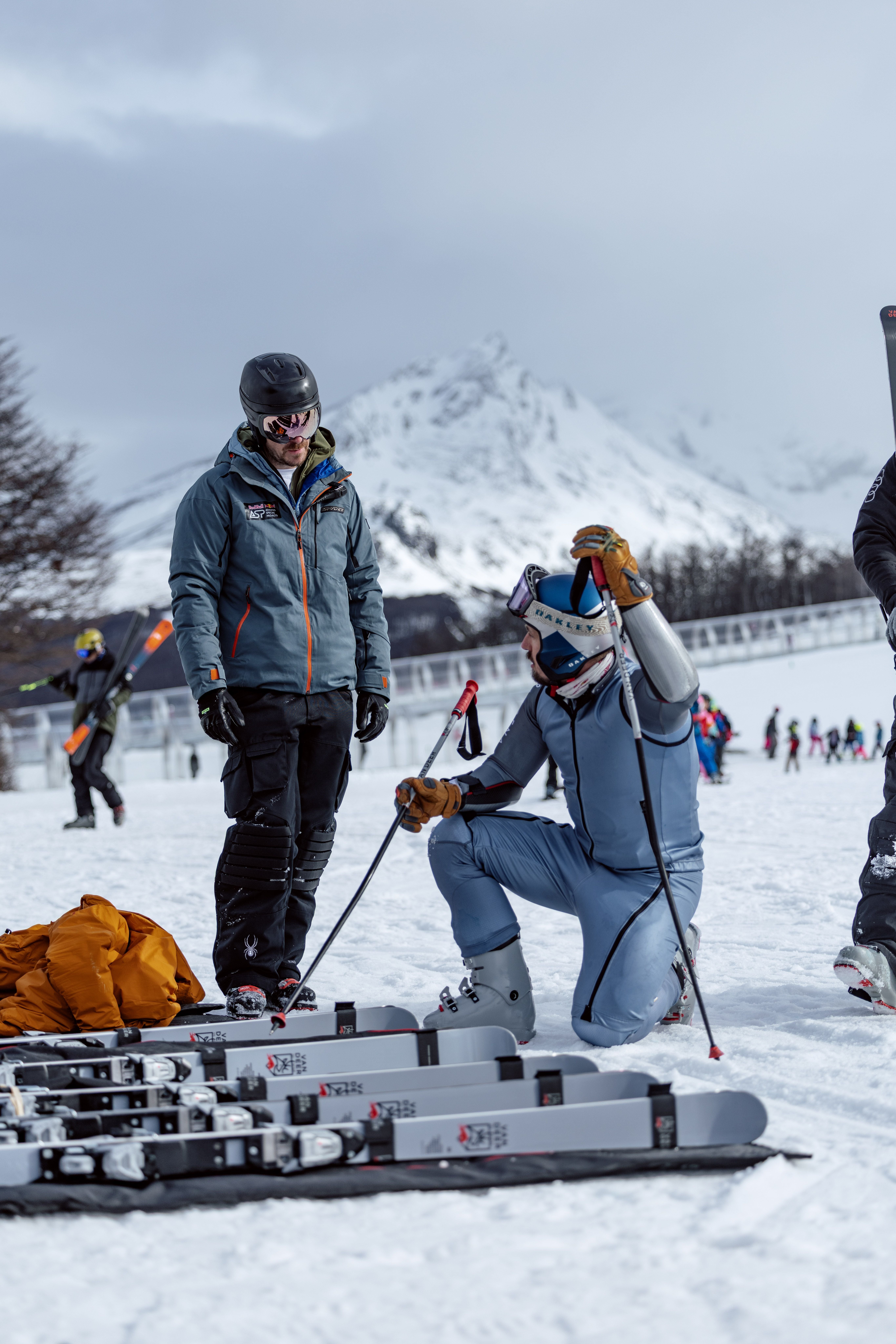 Zwei Skifahrer stehen und knien im Schnee mit Skiern, Berge im Hintergrund.