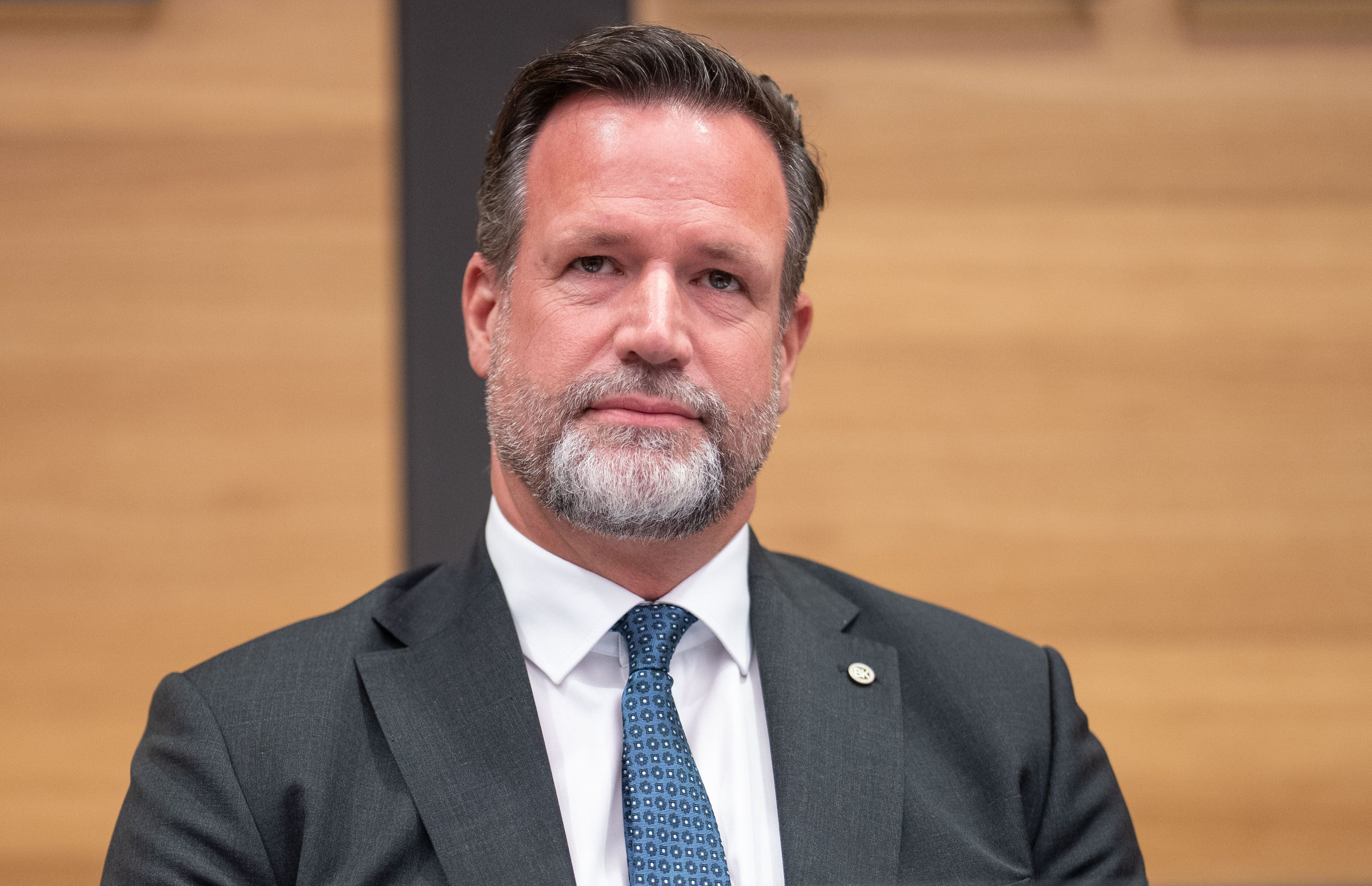 Ein Mann im Anzug mit Krawatte sitzt vor einem holzfarbenen Hintergrund.