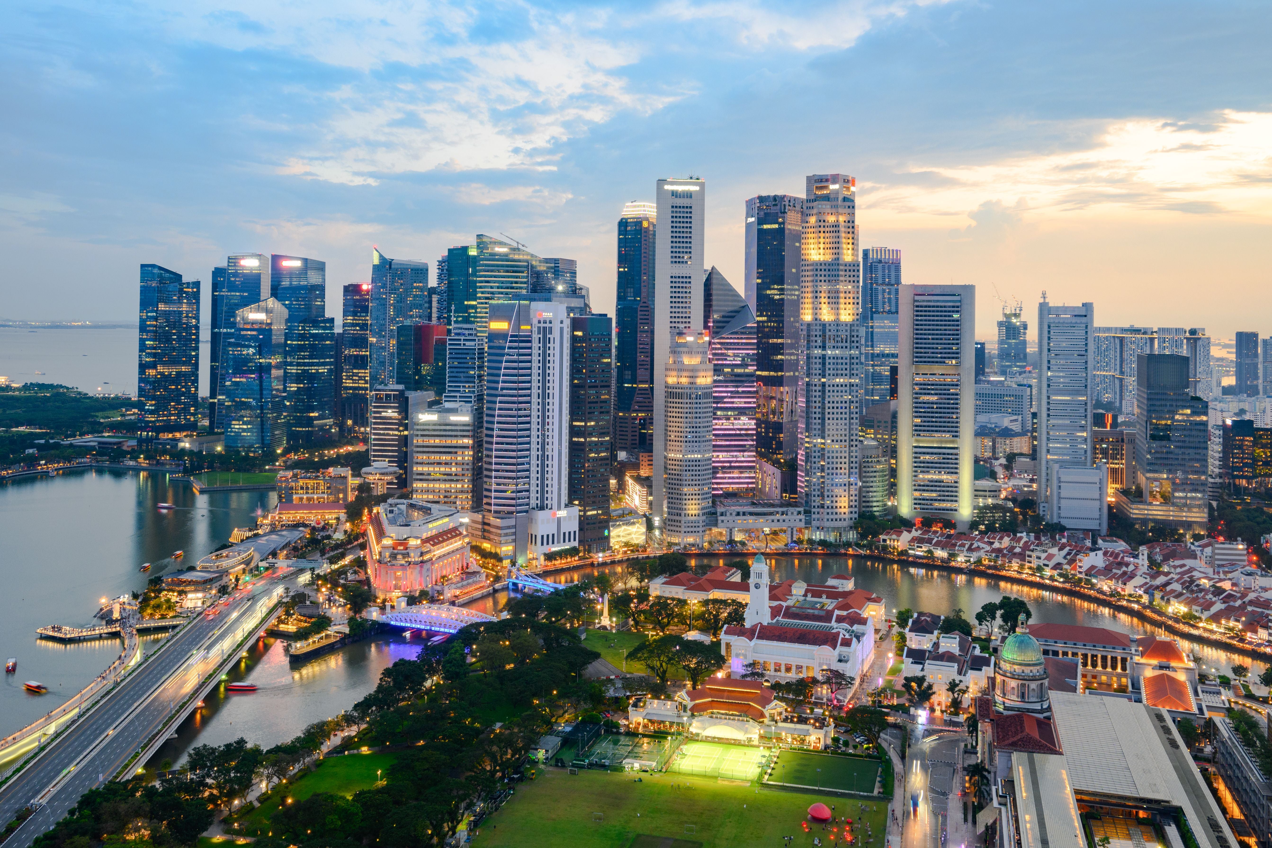 Singapurs Skyline bei Sonnenuntergang, beleuchtete Hochhäuser und Flussufer aus der Luftansicht.