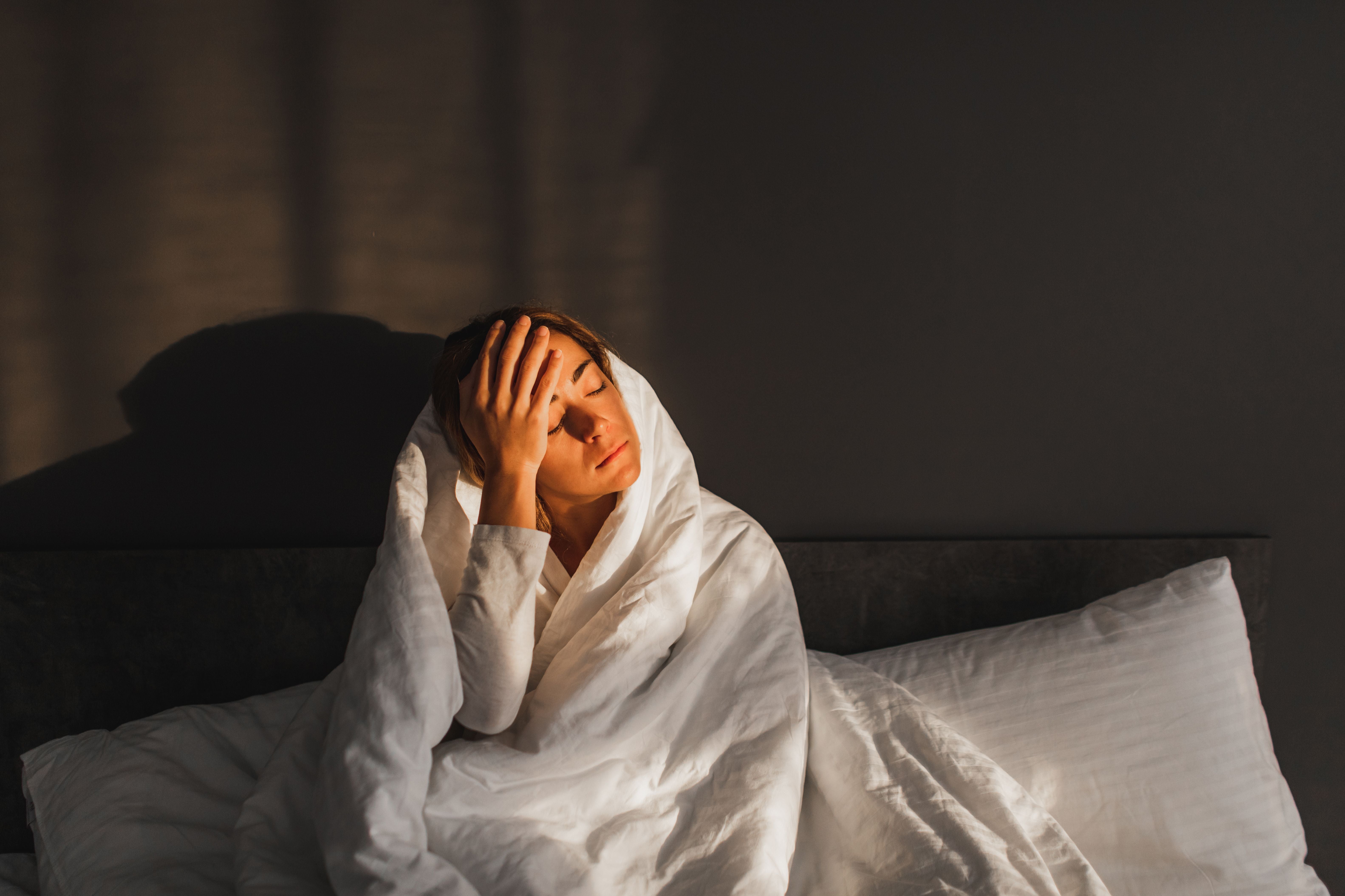 Frau sitzt traurig mit Decke im Bett, hält den Kopf, wirkt erschöpft und krank.