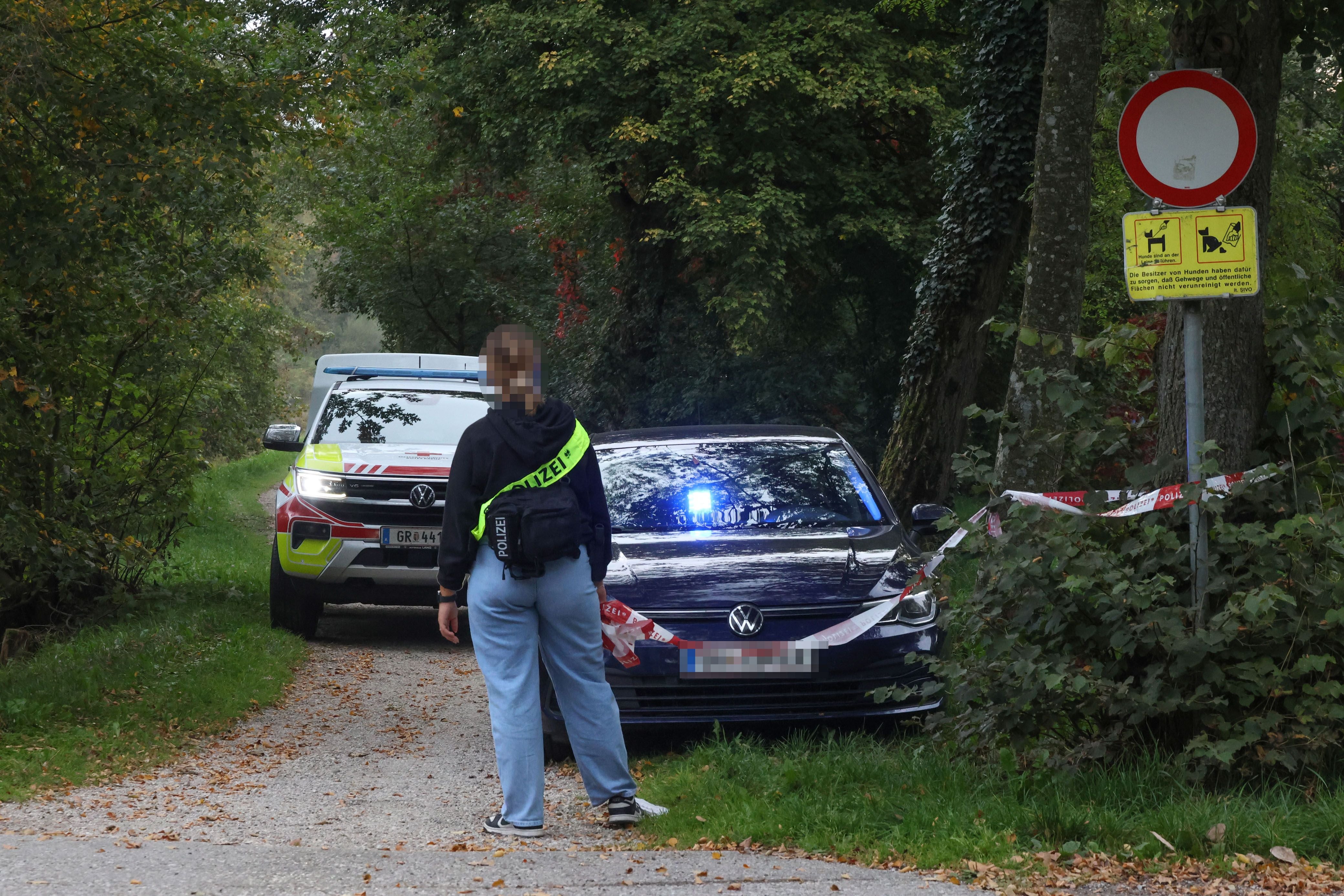 Eine Polizistin steht vor abgesperrtem Weg mit Polizeiautos und Absperrband im Wald.