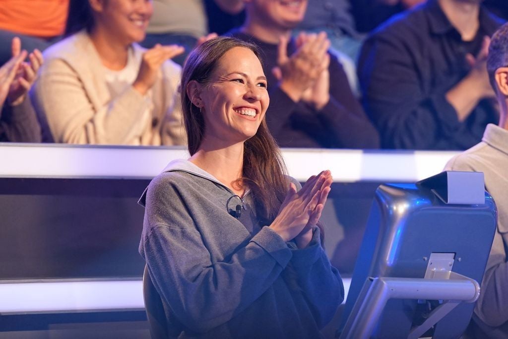Eine Frau sitzt lächelnd in einem Fernsehstudio und klatscht, umgeben von applaudierendem Publikum.