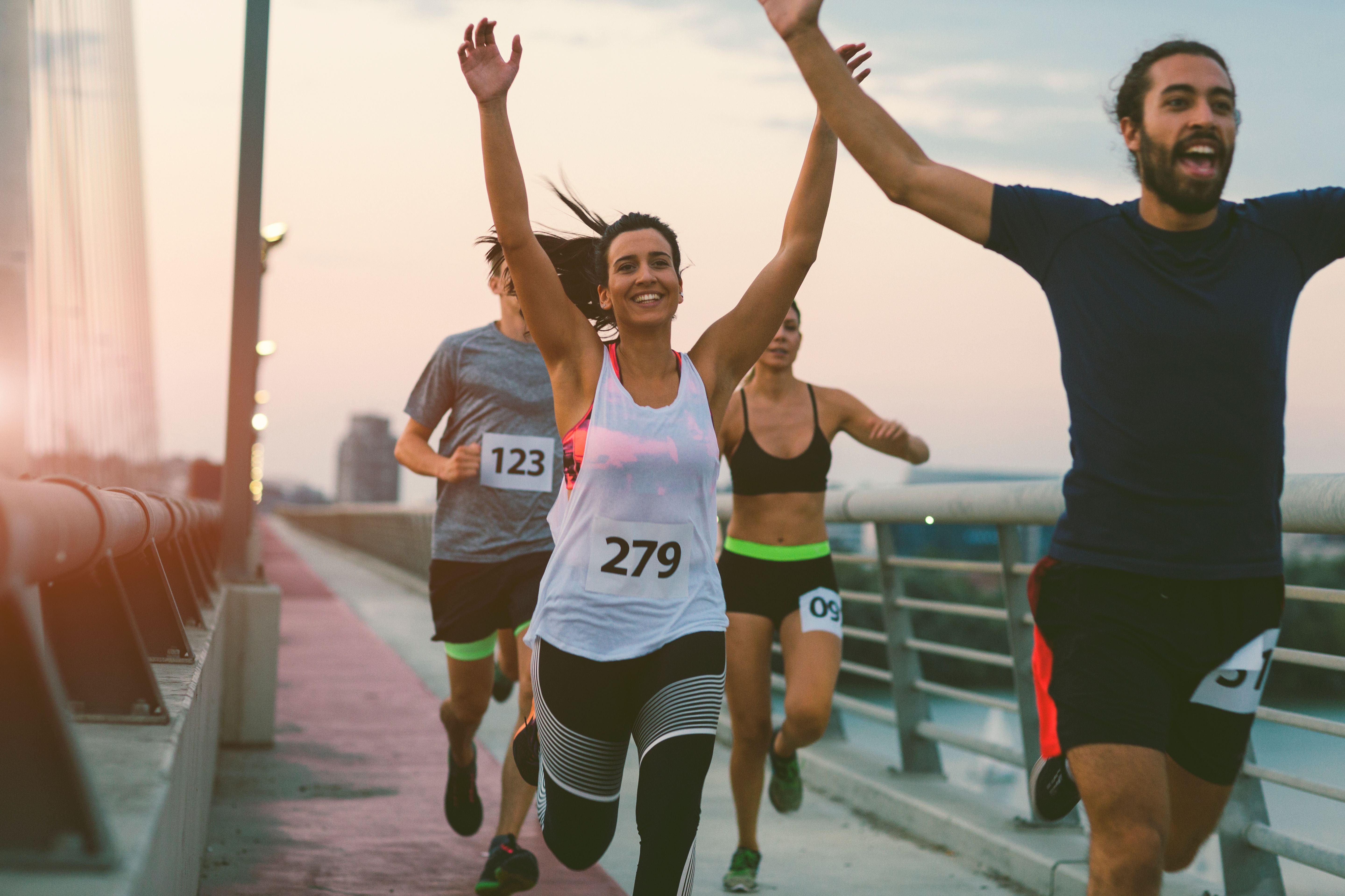 Läufer überqueren jubelnd die Ziellinie eines Marathons auf einer Brücke bei Sonnenuntergang.