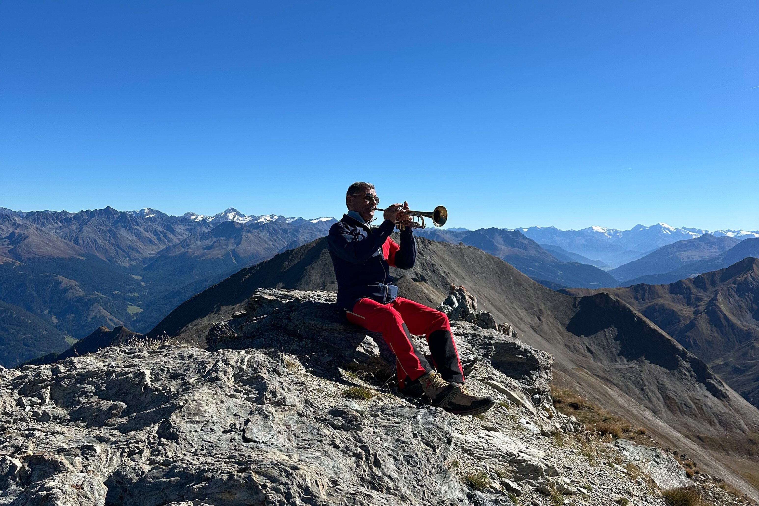 Ein Mann spielt Trompete auf einem Felsen vor beeindruckender Alpen-Bergkulisse bei Serfaus.