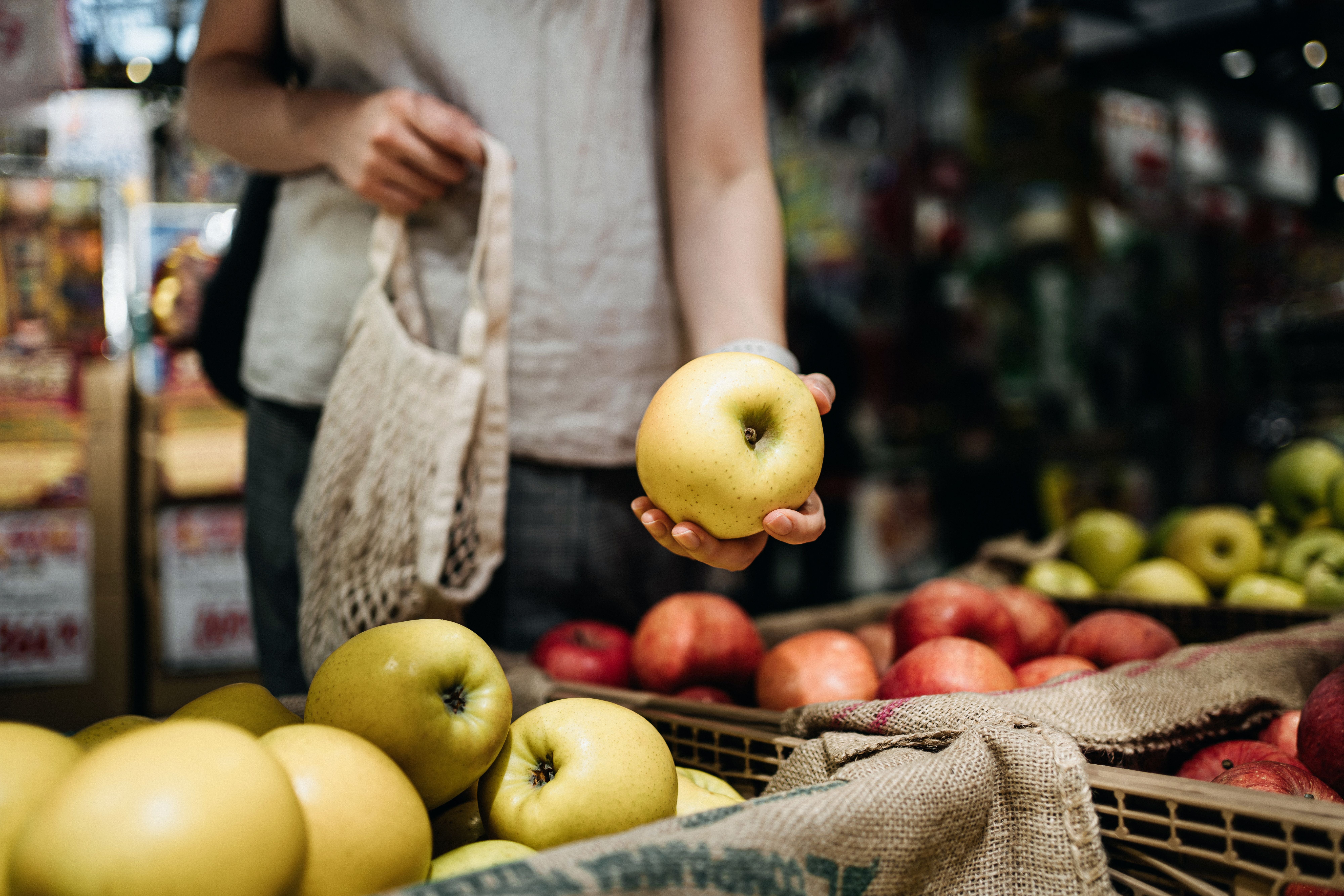 Frau wählt einen gelben Apfel am Obststand und hält eine Baumwolltasche.