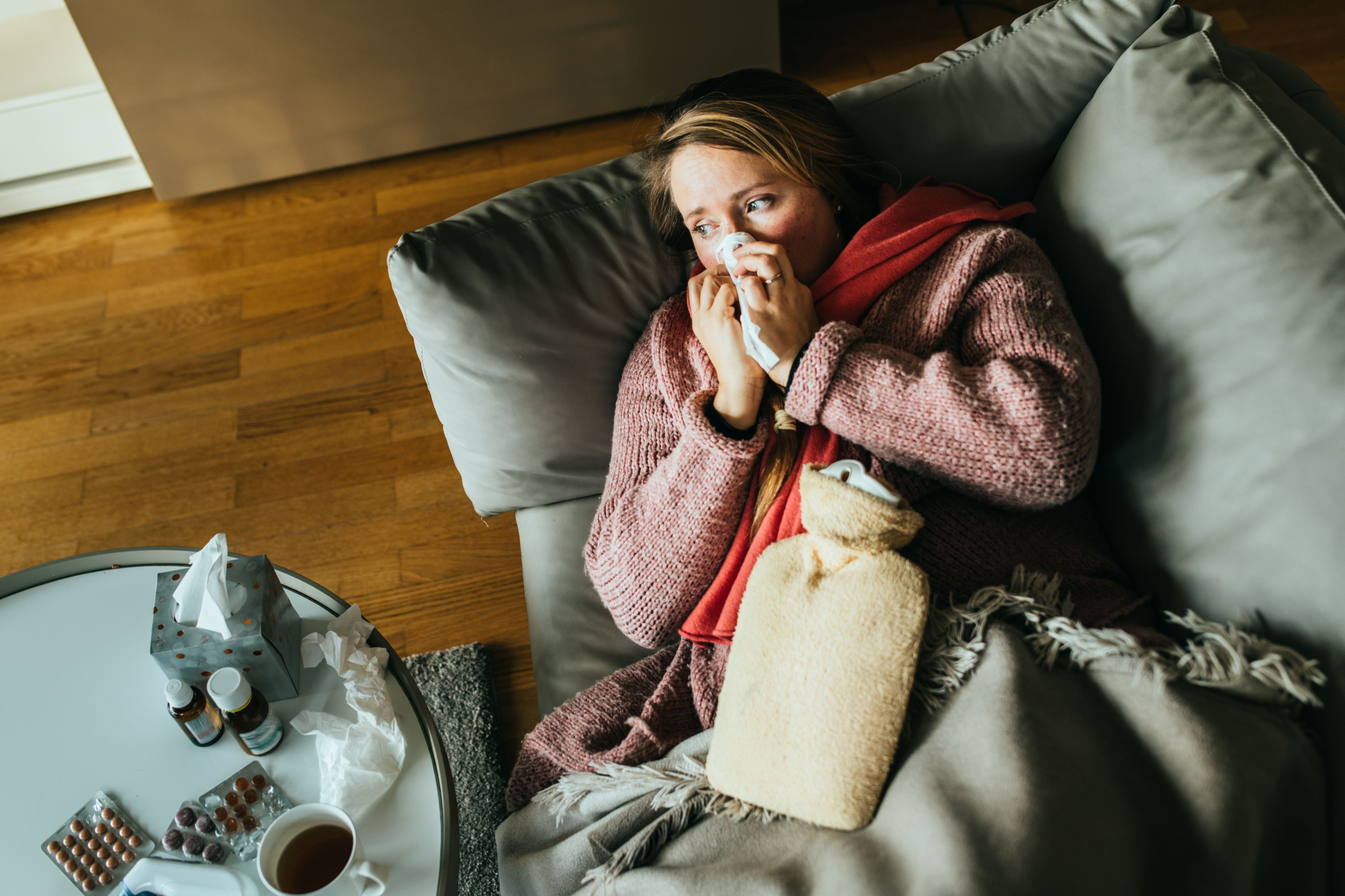 Frau liegt krank mit Wärmflasche auf dem Sofa, Taschentuch am Gesicht, Medikamente auf Tisch.