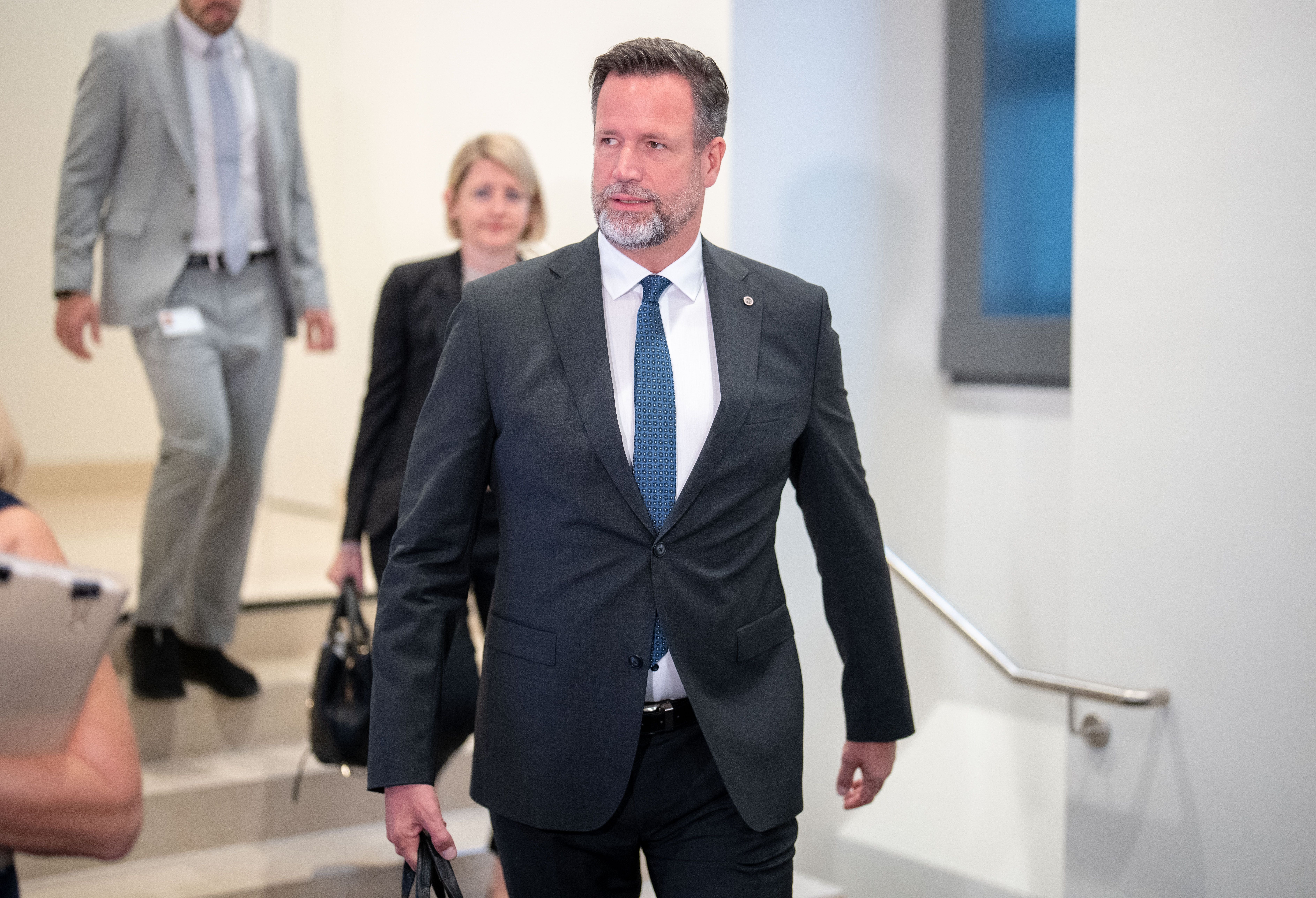 Ein Mann im Anzug geht mit Aktentasche einen Flur entlang, zwei Personen folgen ihm.