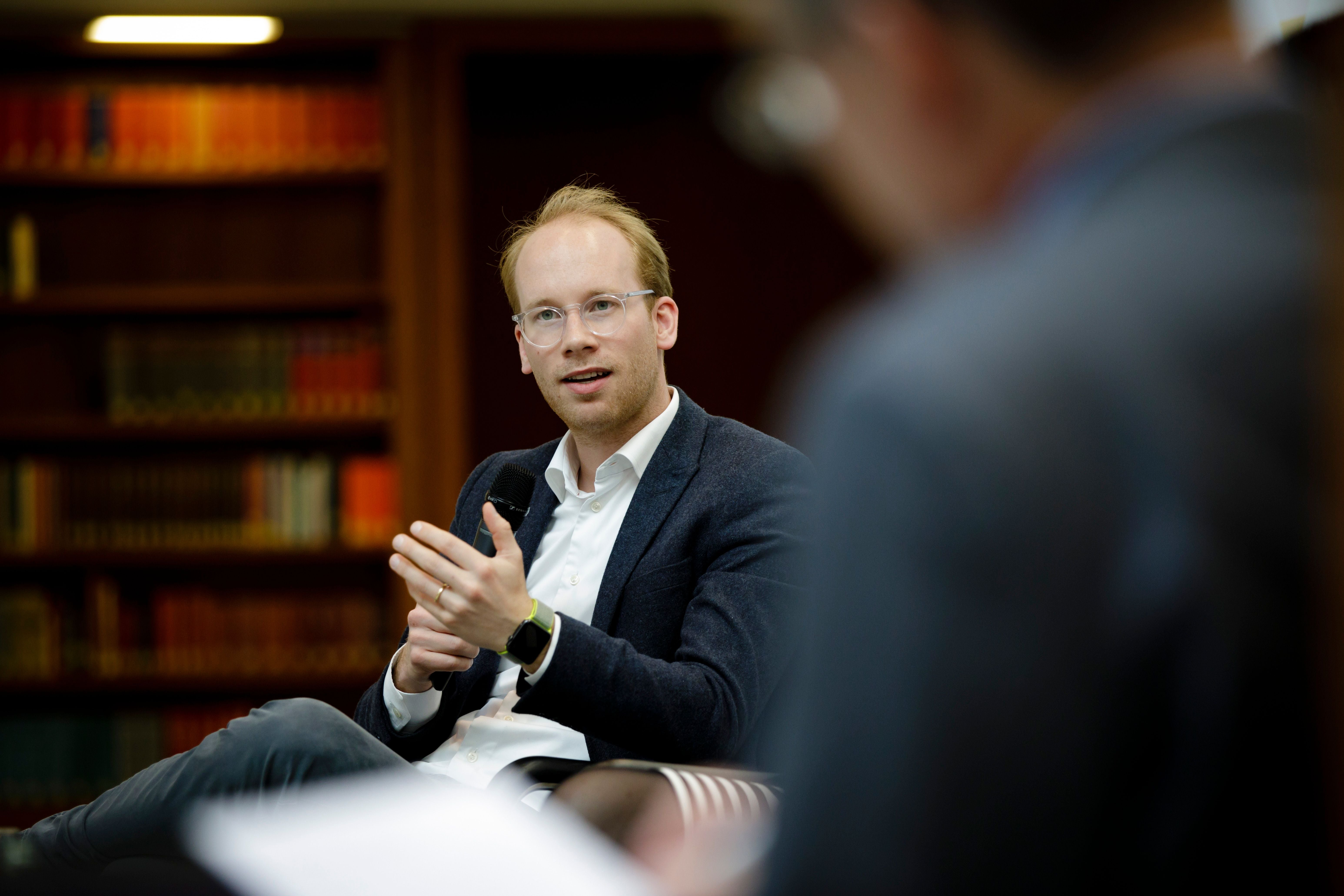 Ein Mann im Gespräch sitzt vor Bücherregalen und gestikuliert mit den Händen.