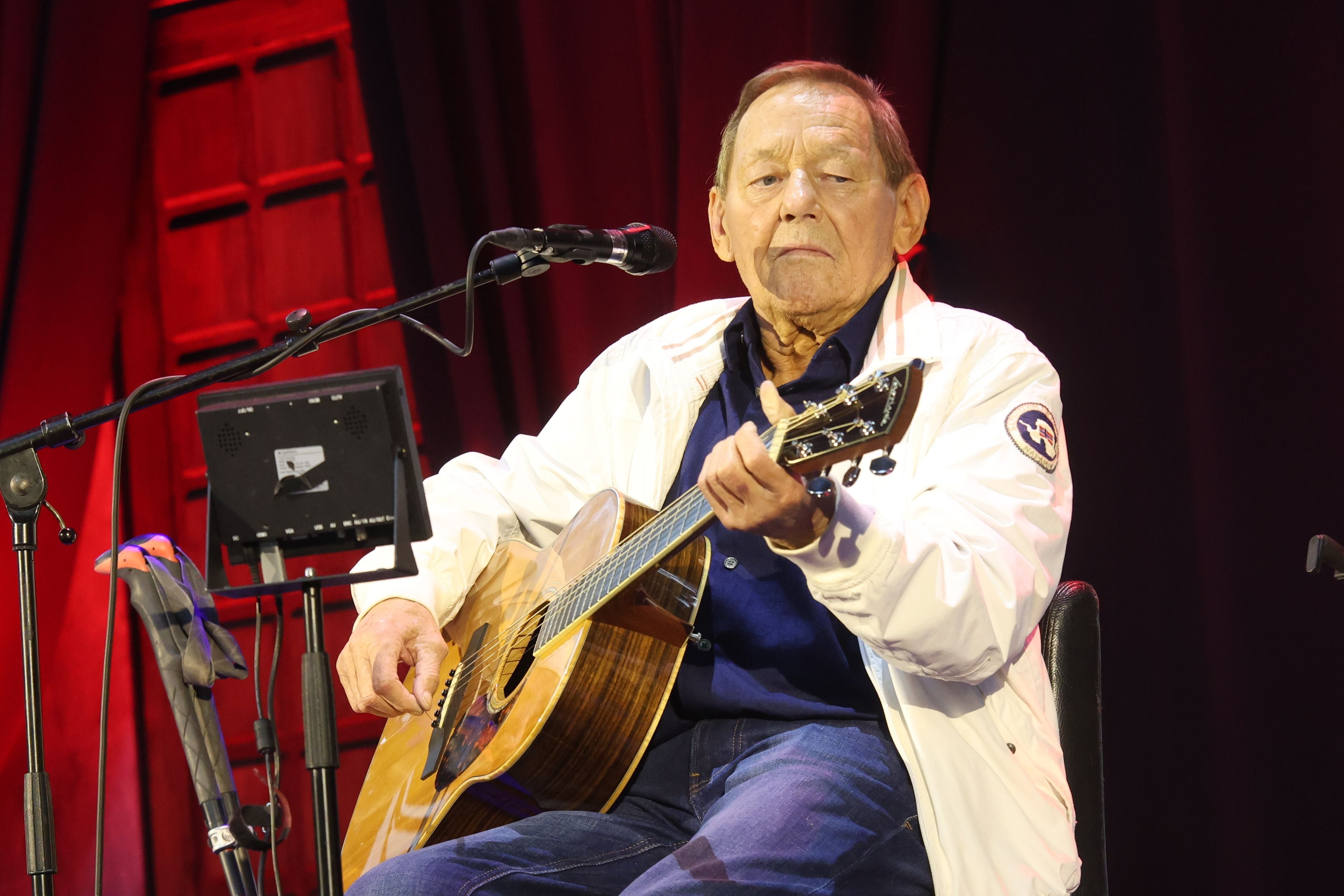 Ein älterer Mann spielt Gitarre auf einer Bühne mit rotem Hintergrund und Mikrofon.