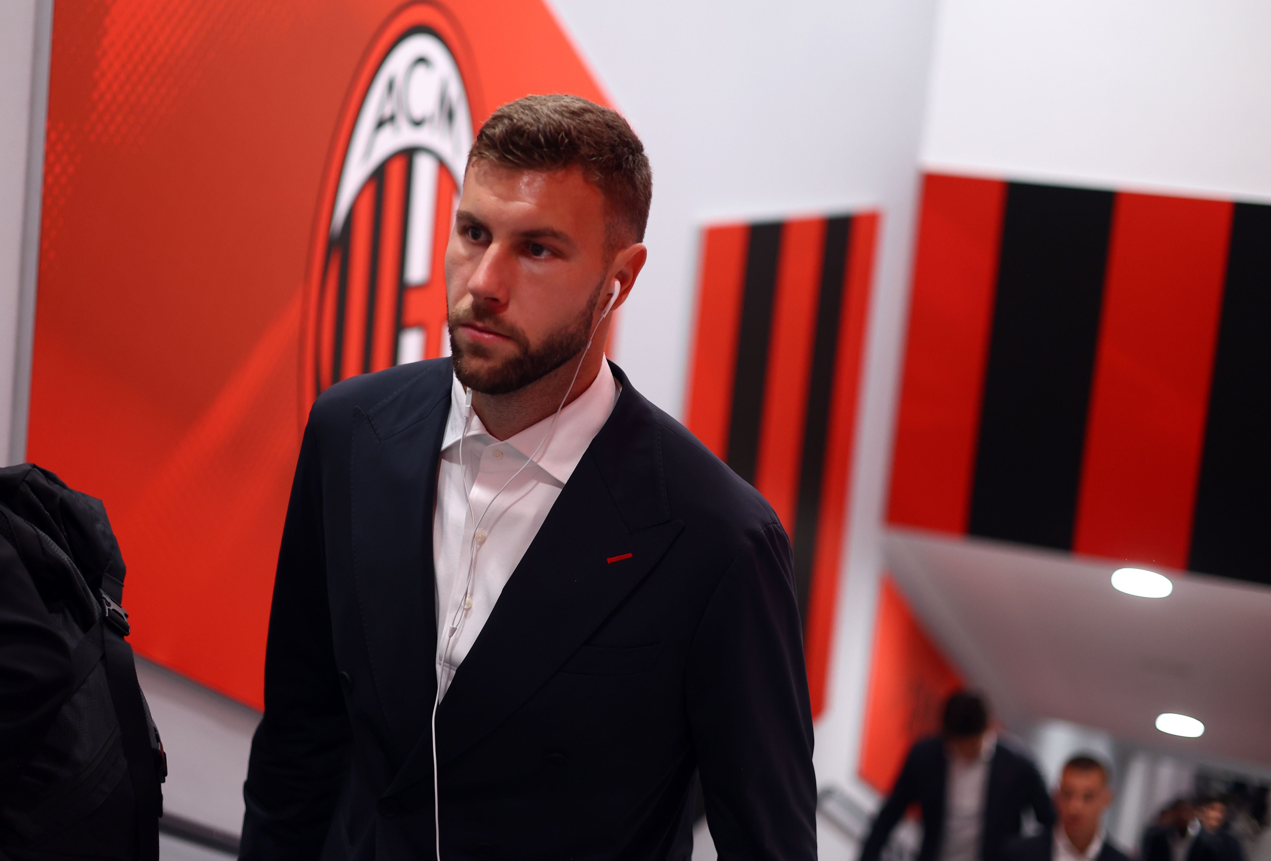 AC-Milan-Profi Strahinja Pavlovic im eleganten dunklen Anzug geht mit Ohrhörern am Vereinswappen der Mailänder vorbei.