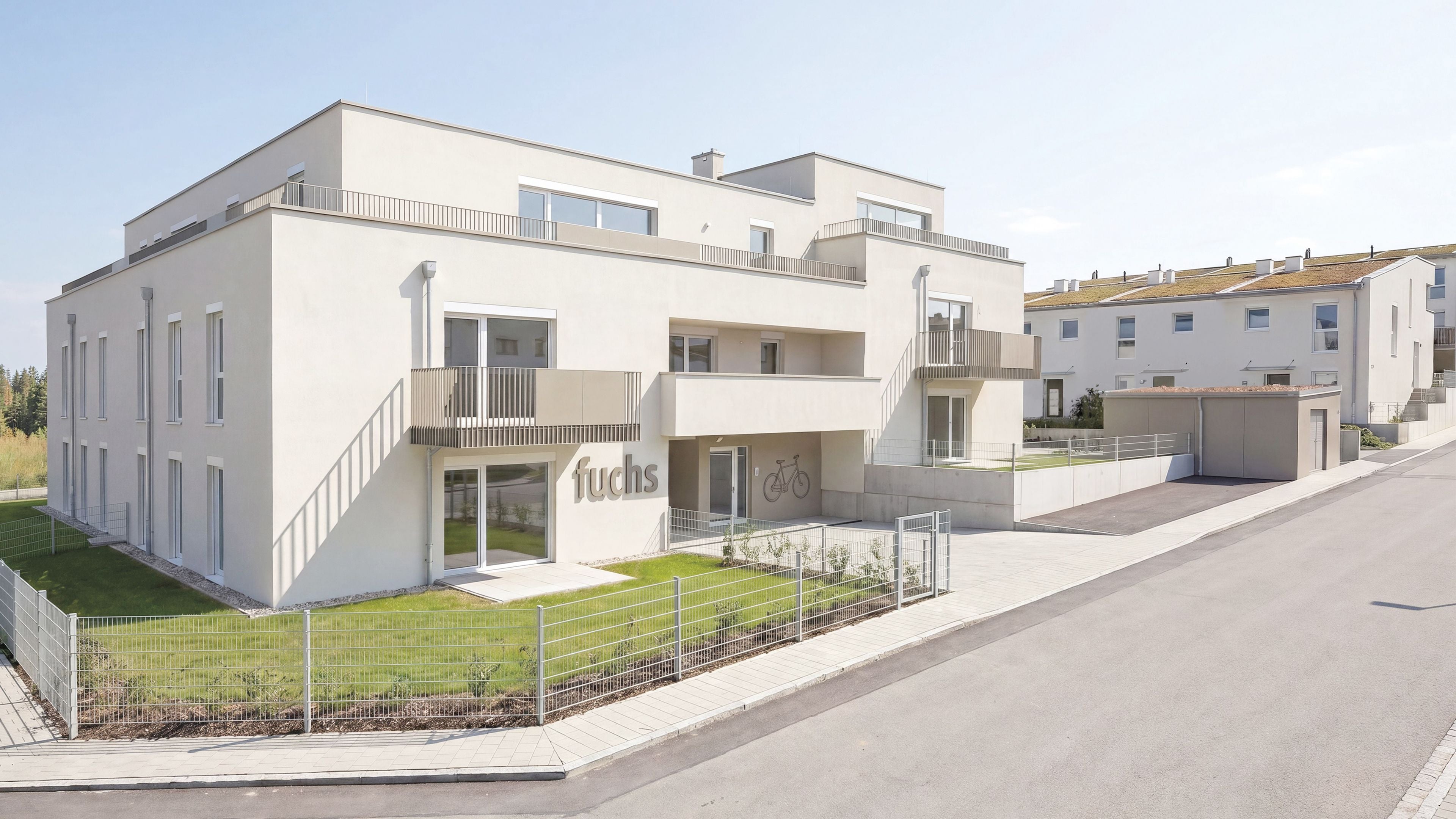 Modernes, weißes Mehrfamilienhaus mit Balkonen und gepflegtem Vorgarten an einer ruhigen Straße.