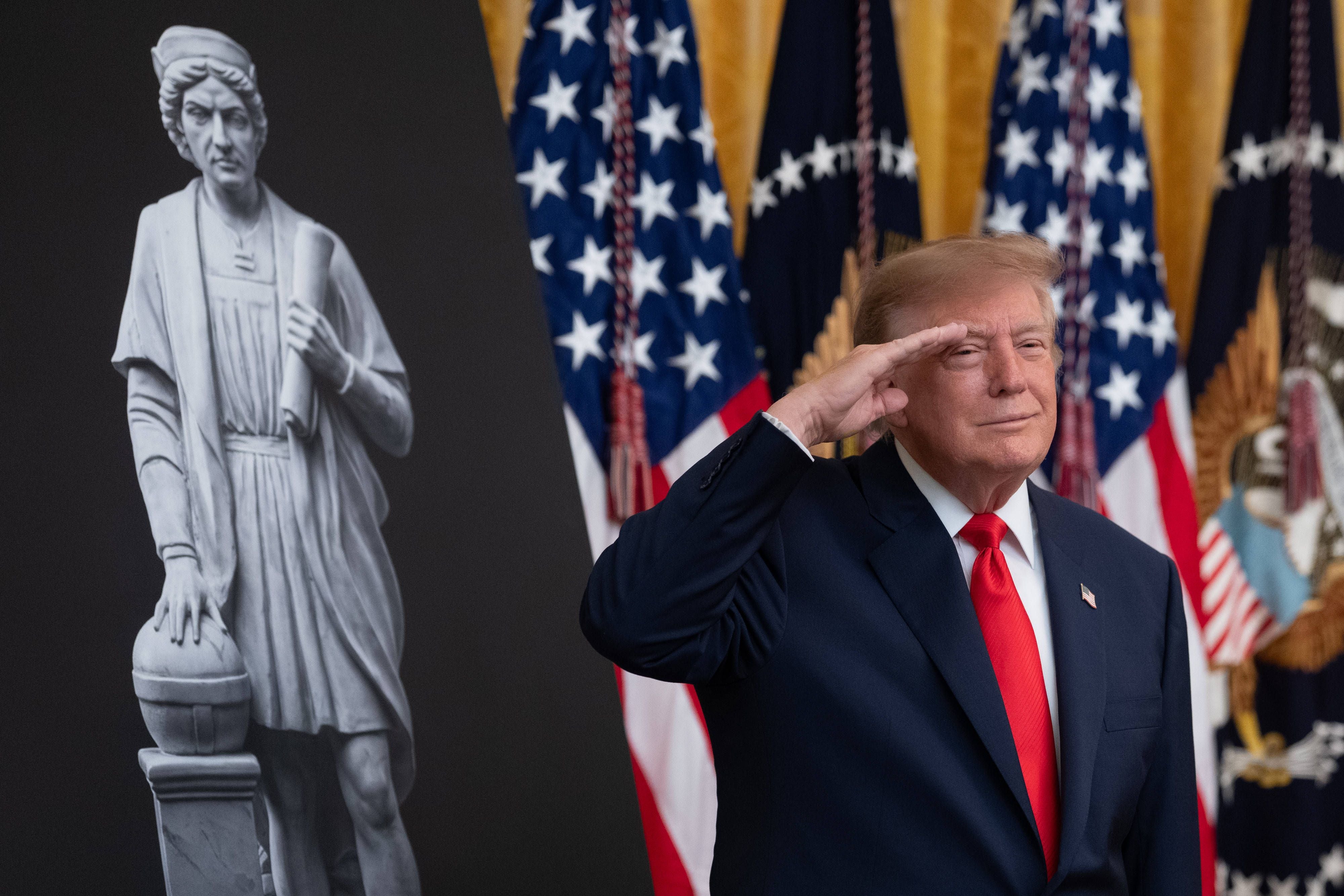 Donald Trump salutiert vor US-Flaggen und einer Kolumbus-Statue im Weißen Haus.