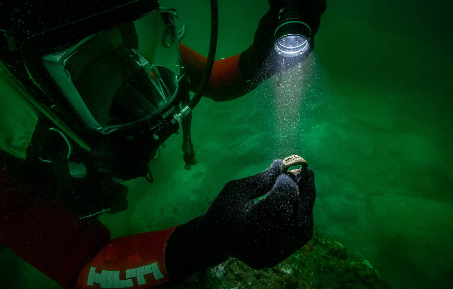 Ein Taucher untersucht unter Wasser mit einer Taschenlampe einen kleinen Fundgegenstand.