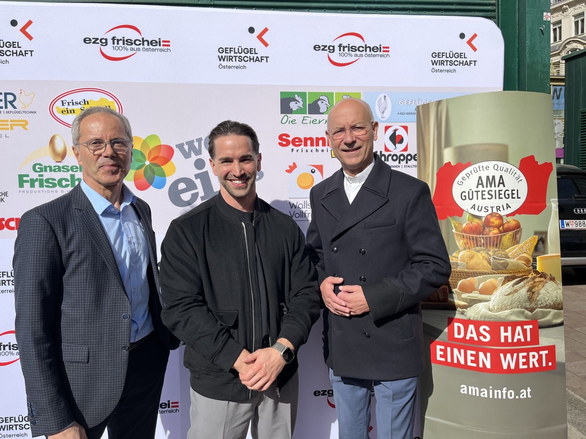 Drei Männer stehen lächelnd bei einer Charity-Veranstaltung am Naschmarkt vor Sponsorenwänden.