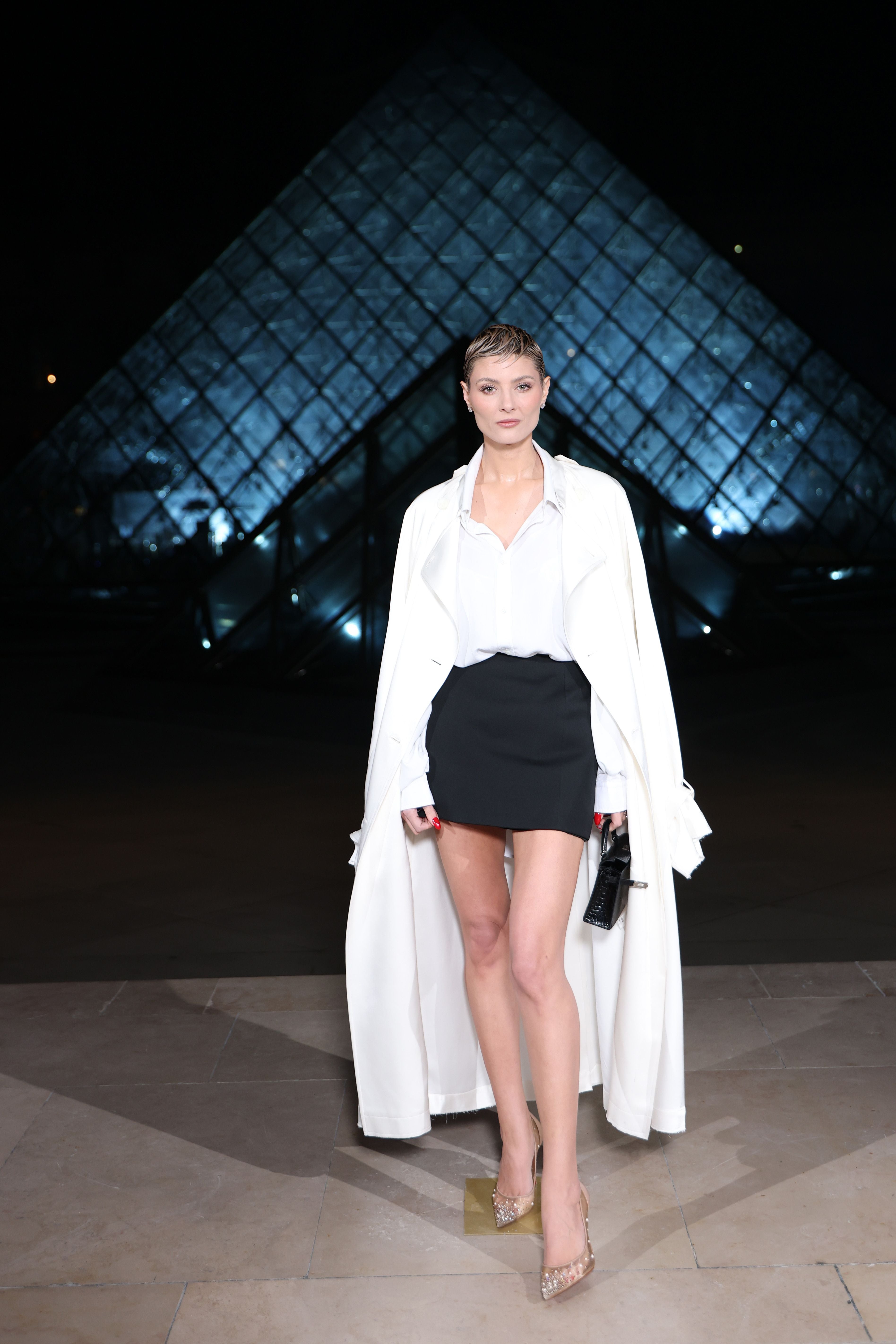 Frau im weißen Mantel vor der beleuchteten Glaspyramide des Louvre bei Nacht.