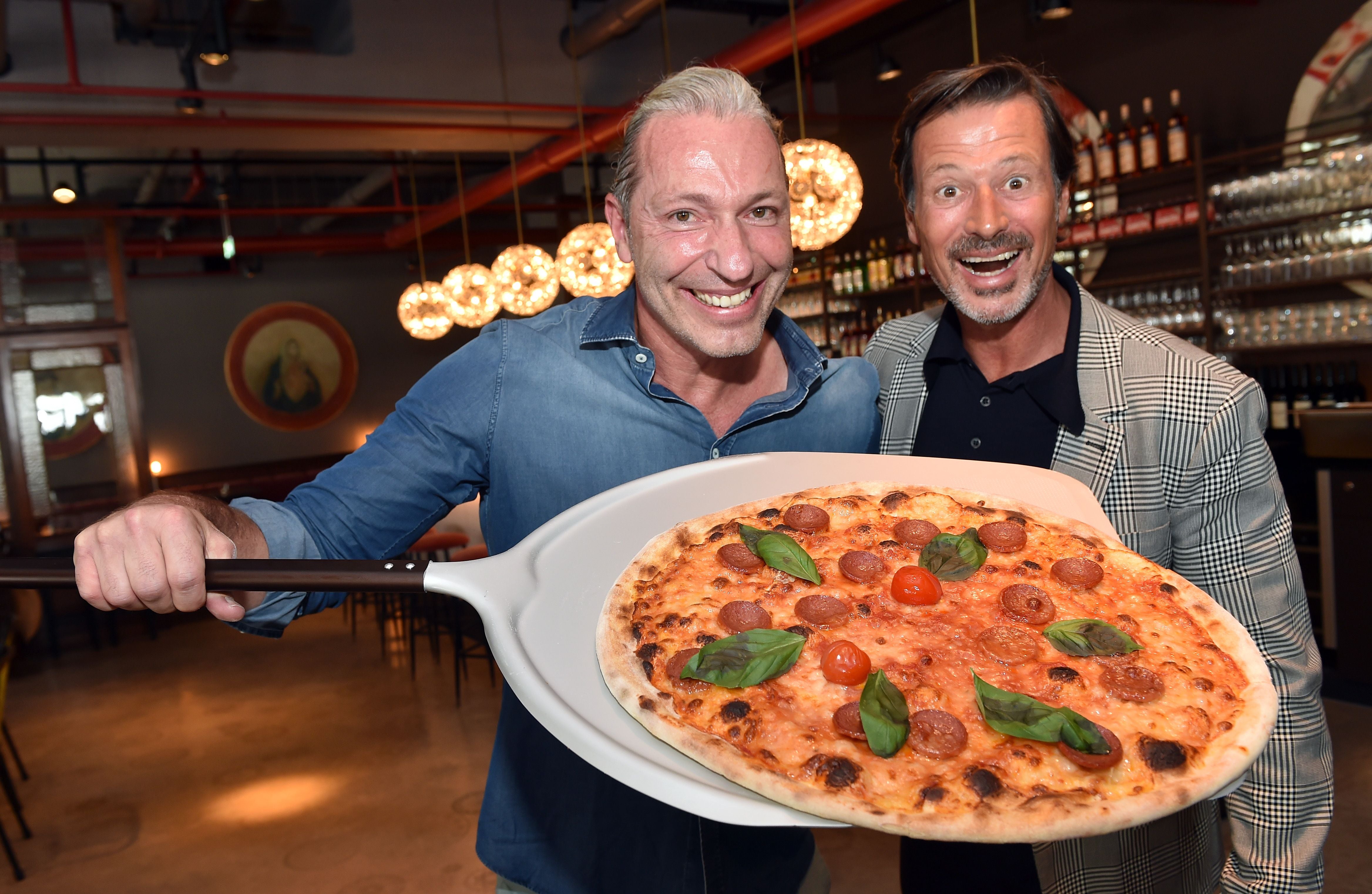 Zwei Männer lächeln und präsentieren in einer Pizzeria eine große Pizza mit Basilikum und Tomaten.