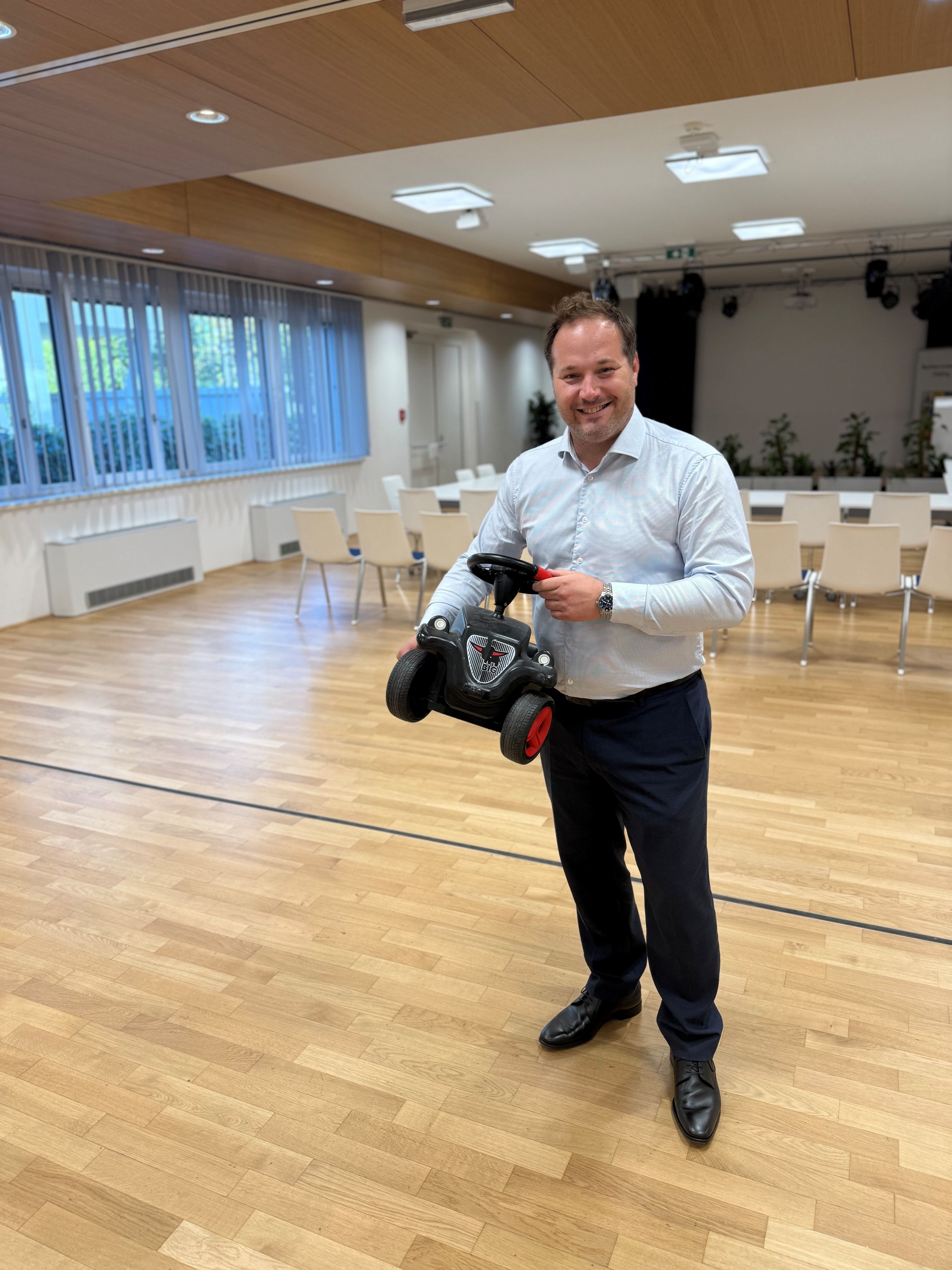 Ein Mann steht in einem Raum und hält ein schwarzes Bobby-Car mit roten Rädern in der Hand.