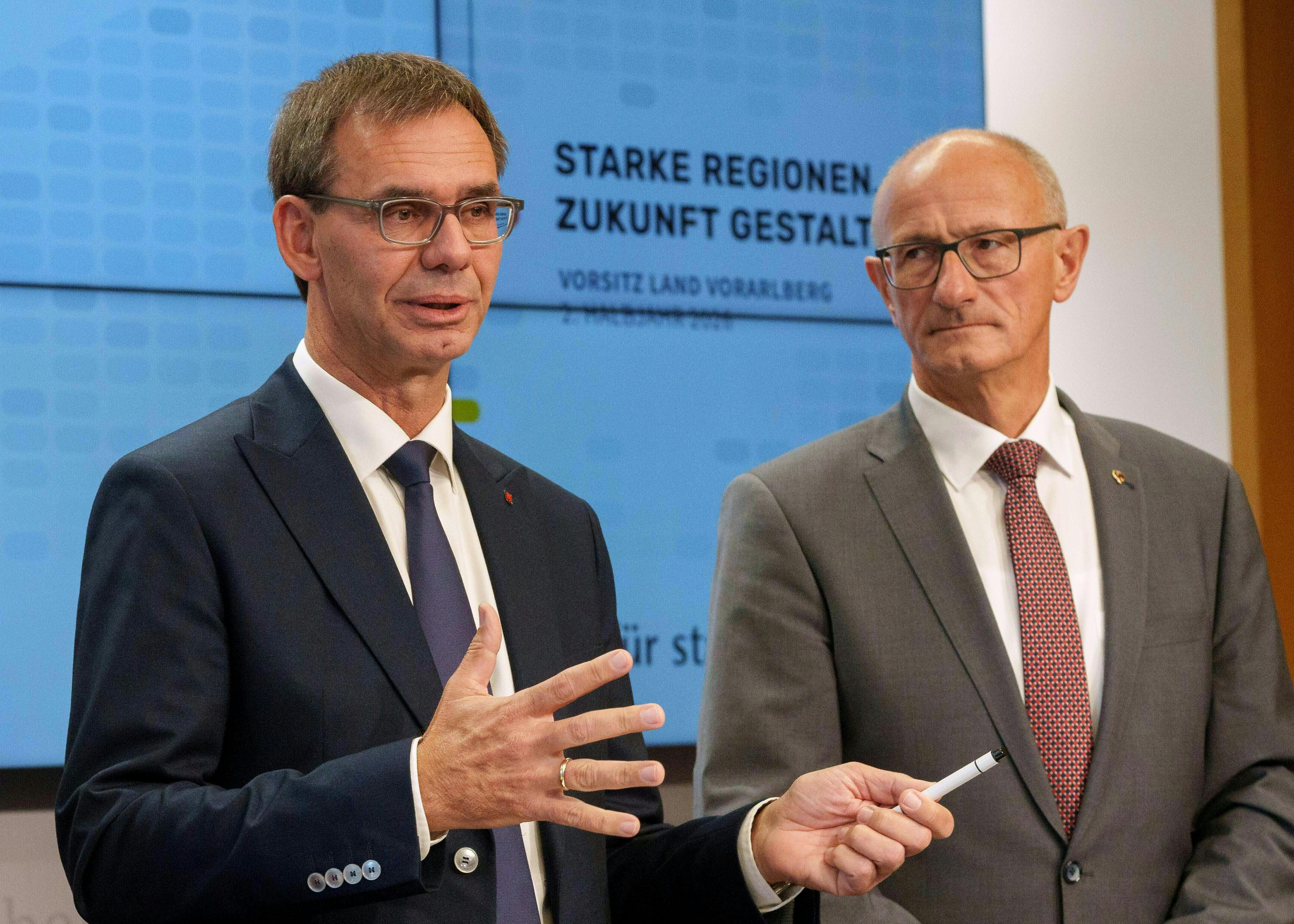 Zwei Männer im Anzug sprechen bei einer Pressekonferenz vor einem Bildschirm.