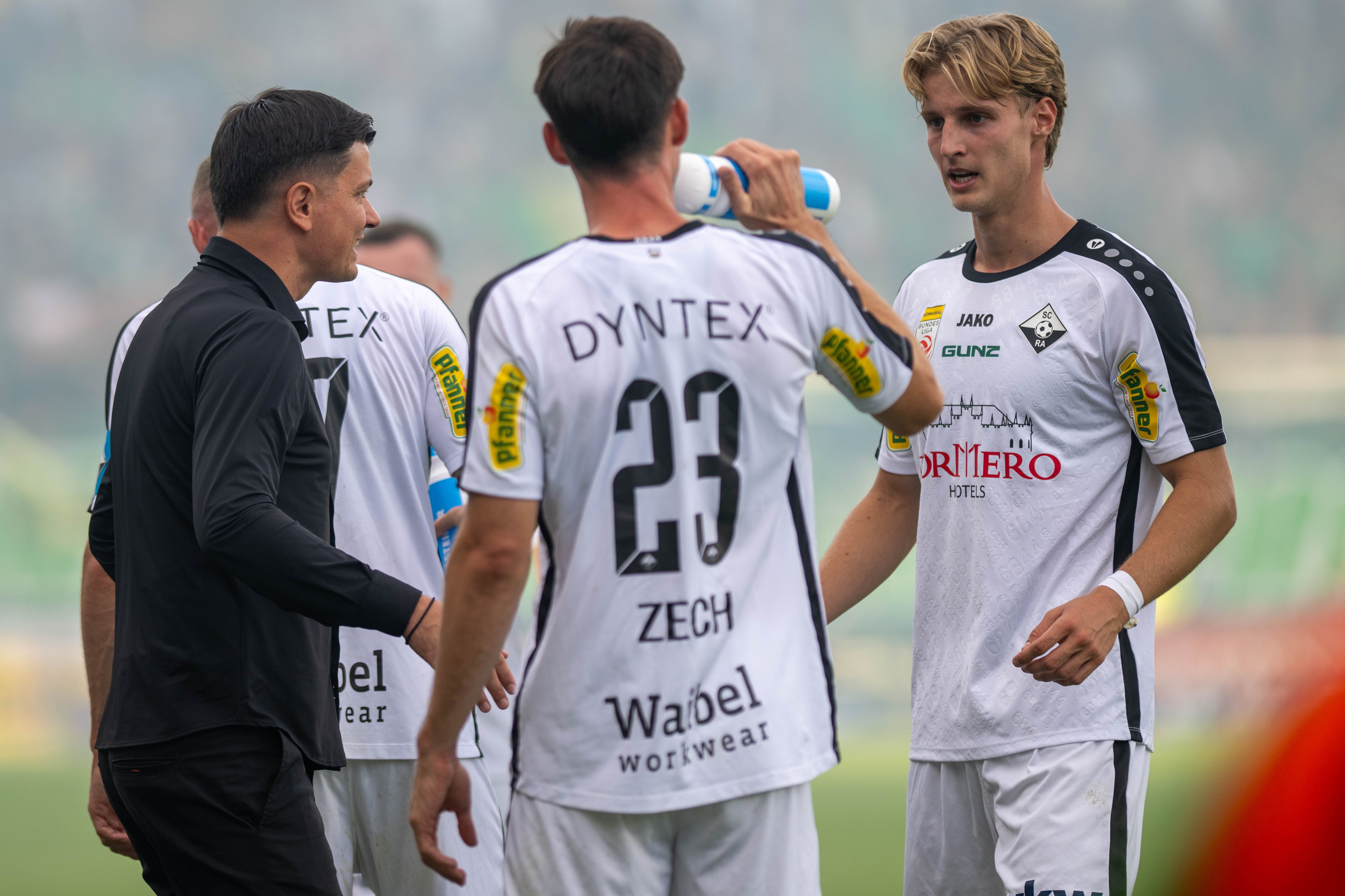 Altach-Trainer Ognjen Zaric spricht am Spielfeldrand intensiv mit zwei Spielern im weißen Trikot, von denen einer aus einer Trinkflasche trinkt.